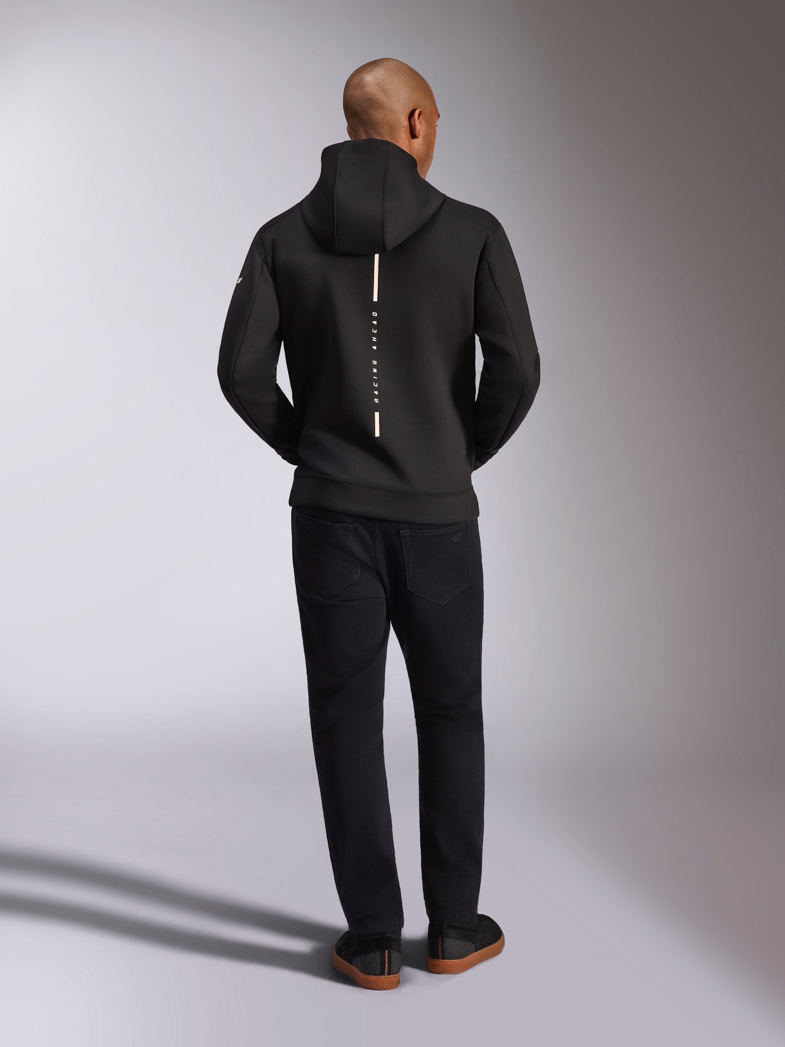 Alpinestars Insight Hoodie, svart, sedd bakifrån på modell, med en vertikal vit randlogotyp på ryggen och texten ”Racing Ahead”, vardaglig huvtröja i tri-blend dubbelsidig fleece, matchad med svarta byxor.