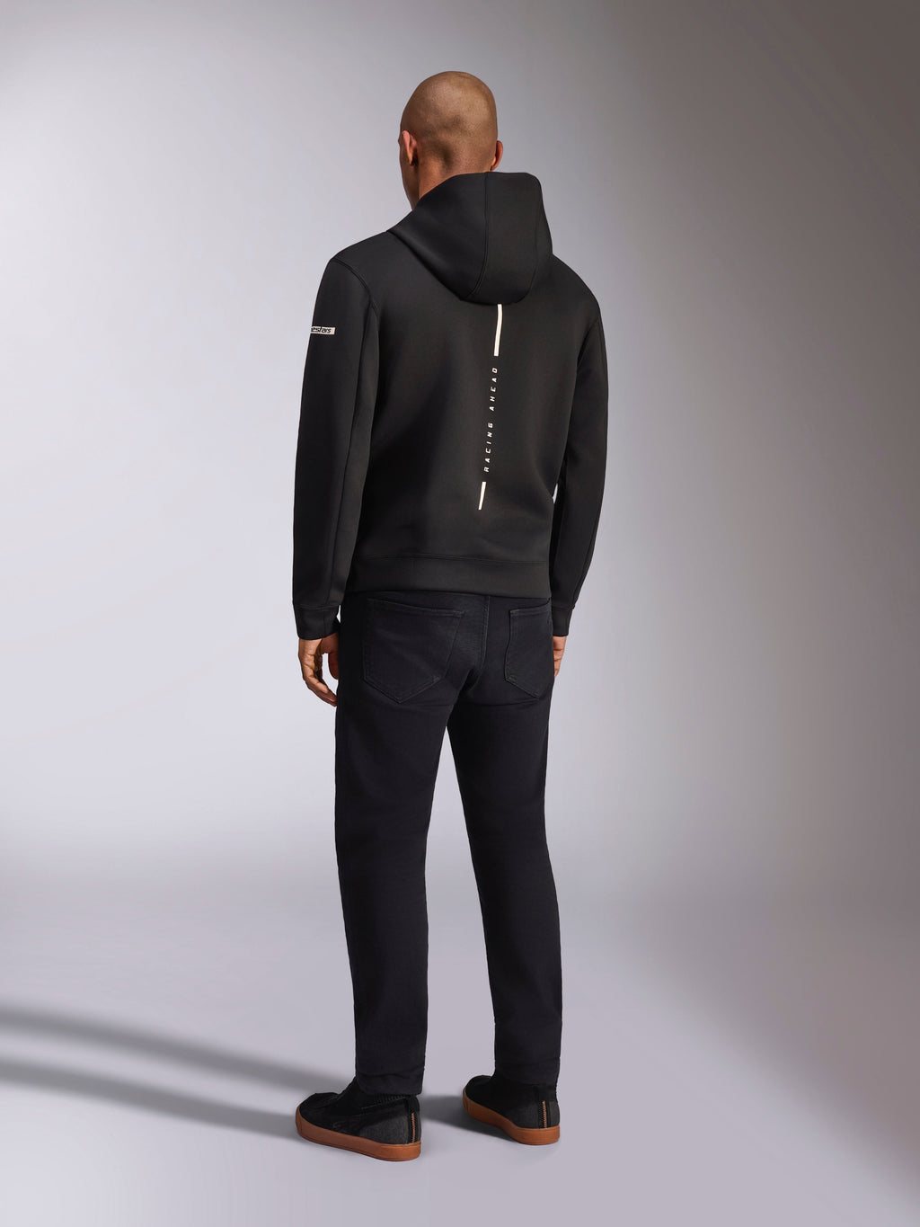 Alpinestars Insight Huvtröja, svart sportswear-huvtröja, vy bakifrån som visar en vertikal vit rand med 'RACING AHEAD'-logotyp, ärmlogotyp, kombinerad med svarta byxor och sneakers, avslappnad motorsportinspirerad design