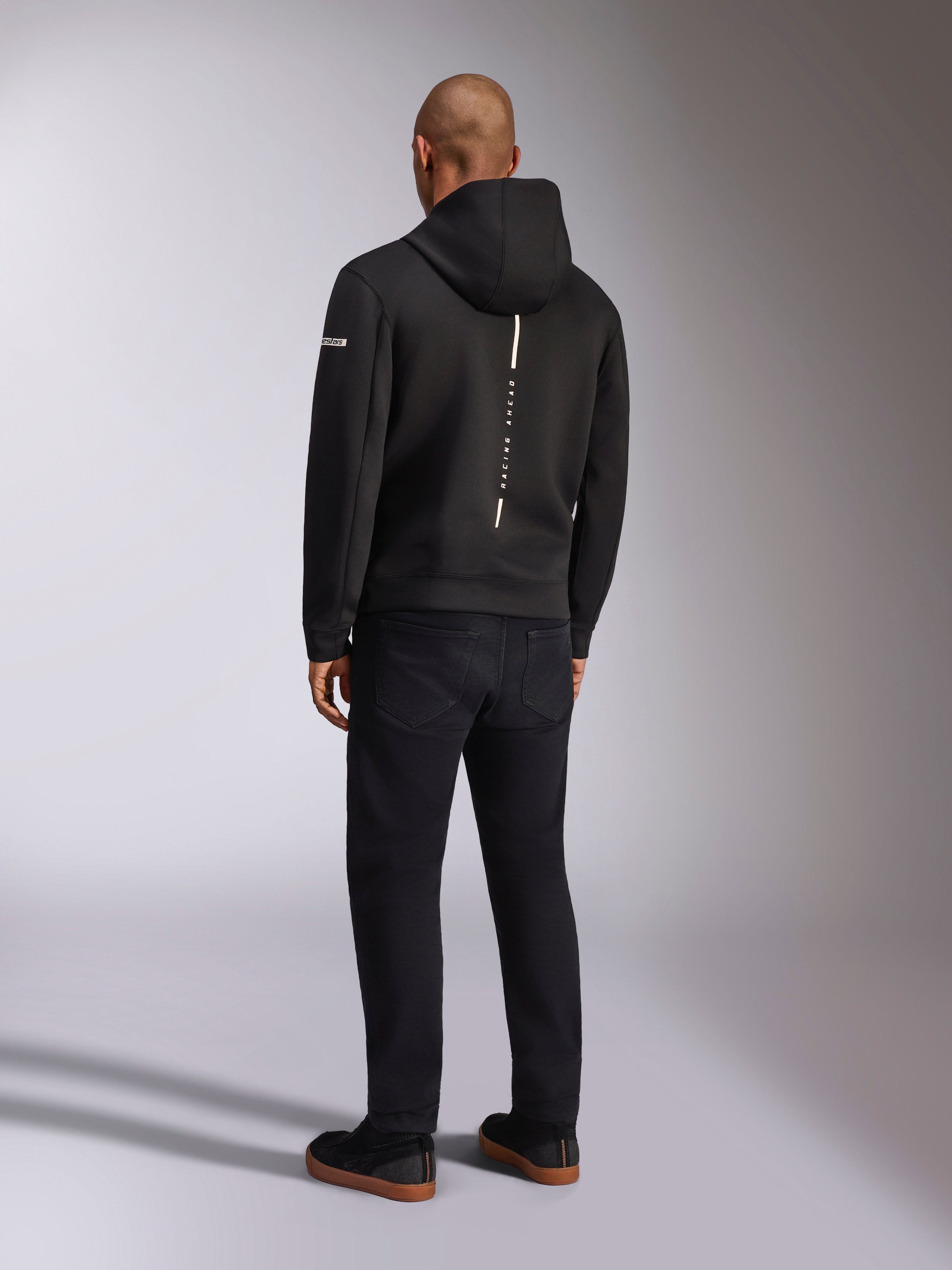 Alpinestars Insight Huvtröja, svart sportswear-huvtröja, vy bakifrån som visar en vertikal vit rand med 'RACING AHEAD'-logotyp, ärmlogotyp, kombinerad med svarta byxor och sneakers, avslappnad motorsportinspirerad design