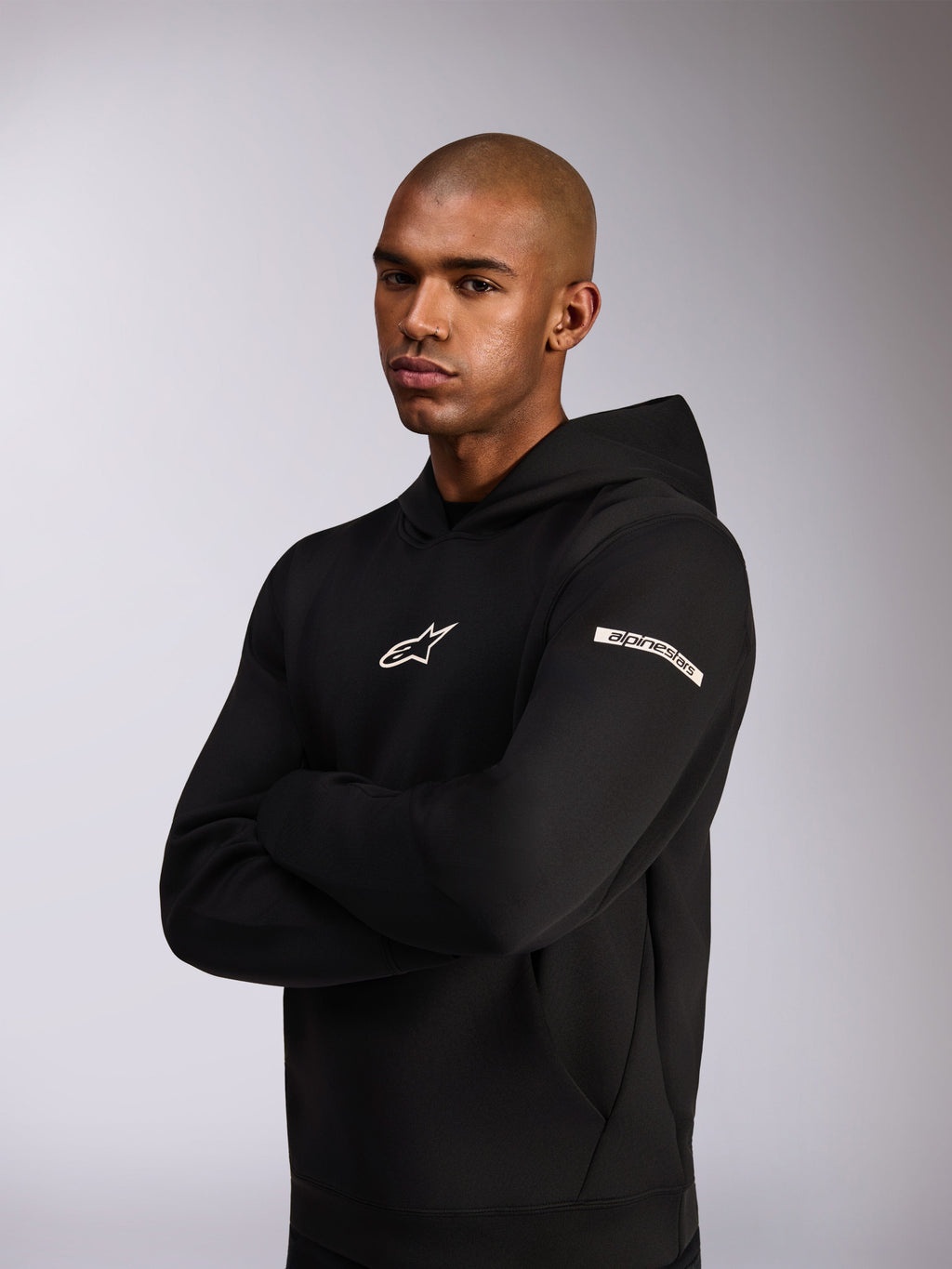 Alpinestars Insight Huvtröja, sportig huvtröja, svart, vit Astars-logotyp på bröstet och ordlogotyp på överarmen, modell som bär huvtröjan med armarna i kors, premiumkonstruktion i dubbelsidig fleece