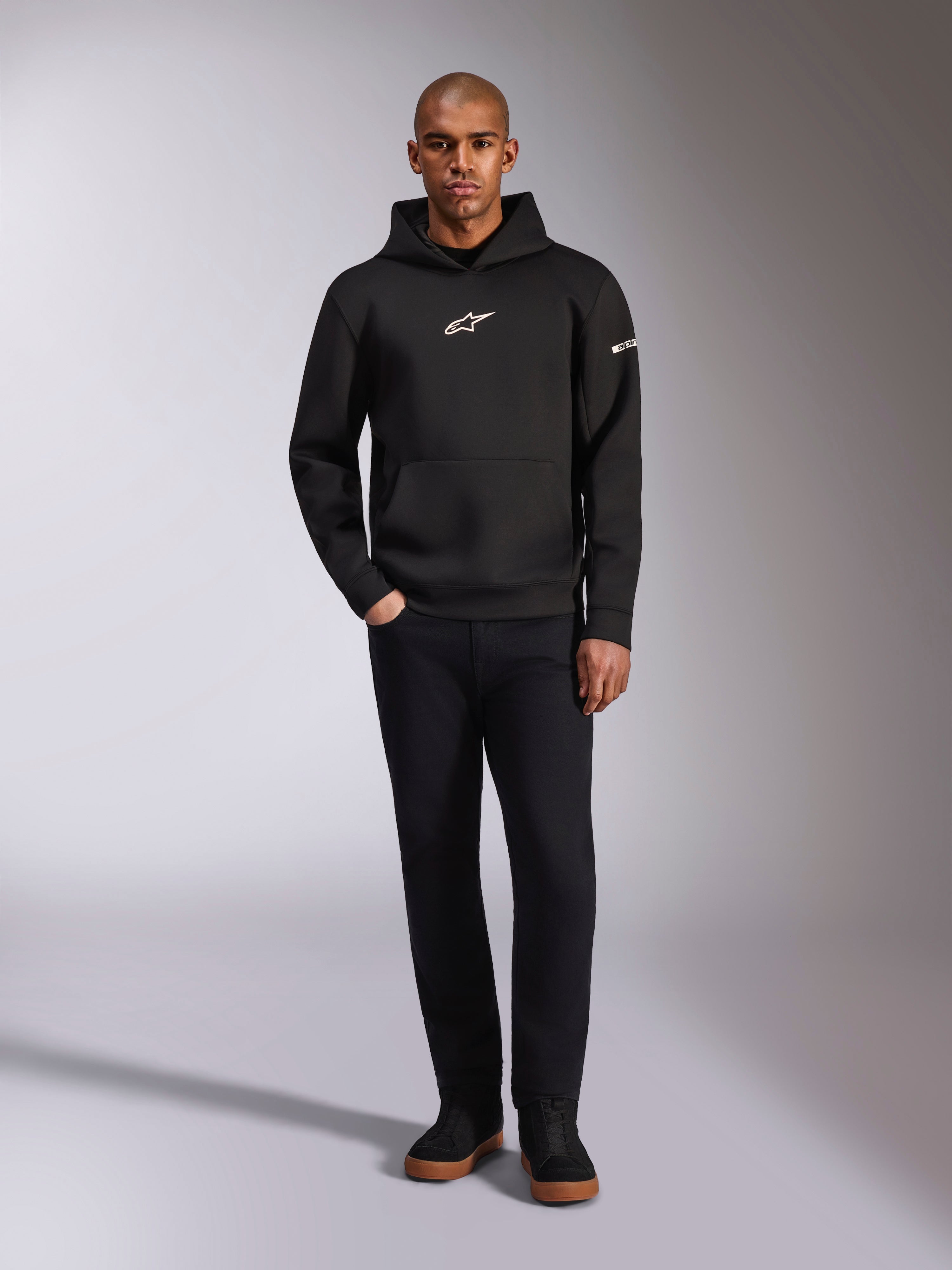 Alpinestars Insight Hoodie, svart huvtröja med vit Astars-logotyp på bröstet och ärmen, bärs av en modell med svarta byxor och svarta sneakers, avslappnad motorsportinspirerad streetwear