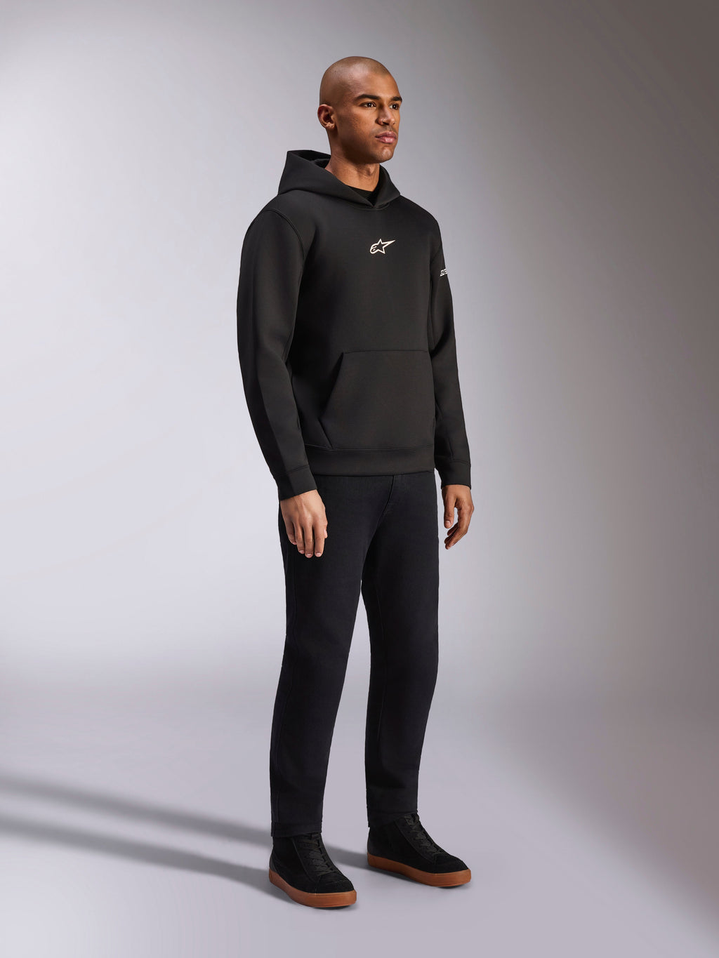 Alpinestars Insight Hoodie, svart, ledig huvtröja i fleece med vit Astars-logotyp på bröstet, känguruficka, bärs med svarta byxor och sneakers, studiobild i helfigur, motorsportinspirerad streetwear