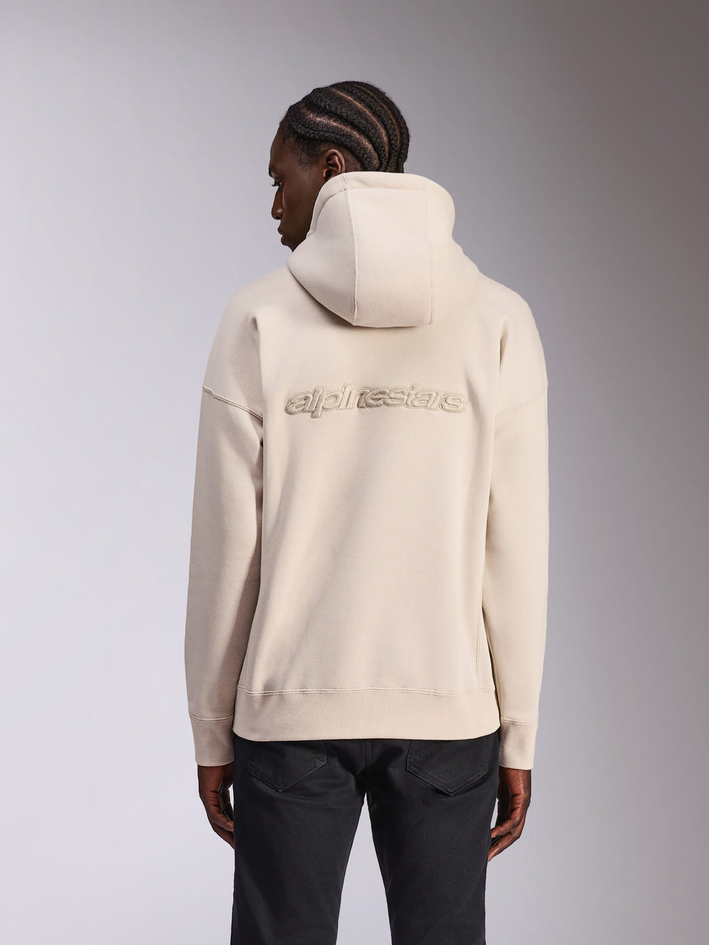 Alpinestars Emergent Oversized Hoodie, casual streetwear, sandfärgad off-white, med 3D-broderad Alpinestars-ordlogotyp på ryggen, oversized passform med ribbad fåll och muddar, vy bakifrån på modell som bär svarta byxor.