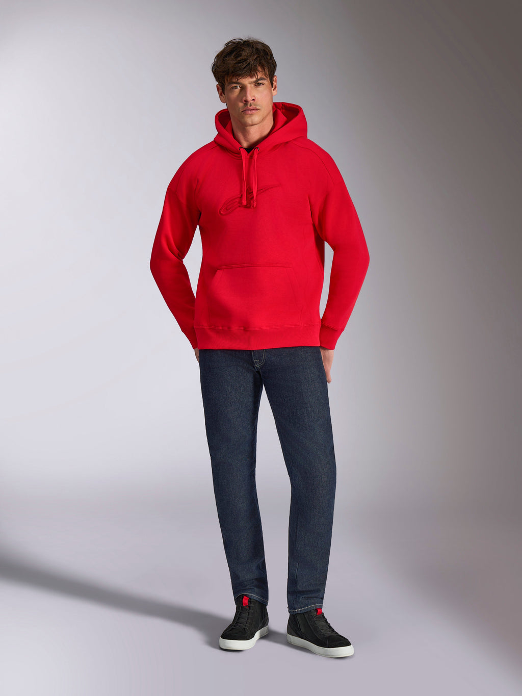 Alpinestars Emergent Oversized Hoodie, röd, med ton-i-ton 3D-broderad Astars-logotyp på bröstet, känguruficka och huva med dragsnöre, bärs av en modell med mörkblå jeans och svarta sneakers, avslappnad lifestyle-streetwear