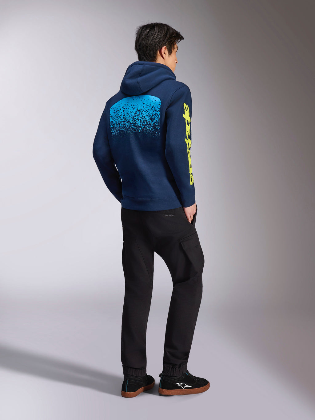 Alpinestars Obsessed Hoodie, marinblå sweatshirt, vy bakifrån som visar ljusblå grafik med MX-gradering och gul Alpinestars-logotyp på ärmen, bärs med svarta byxor och sneakers