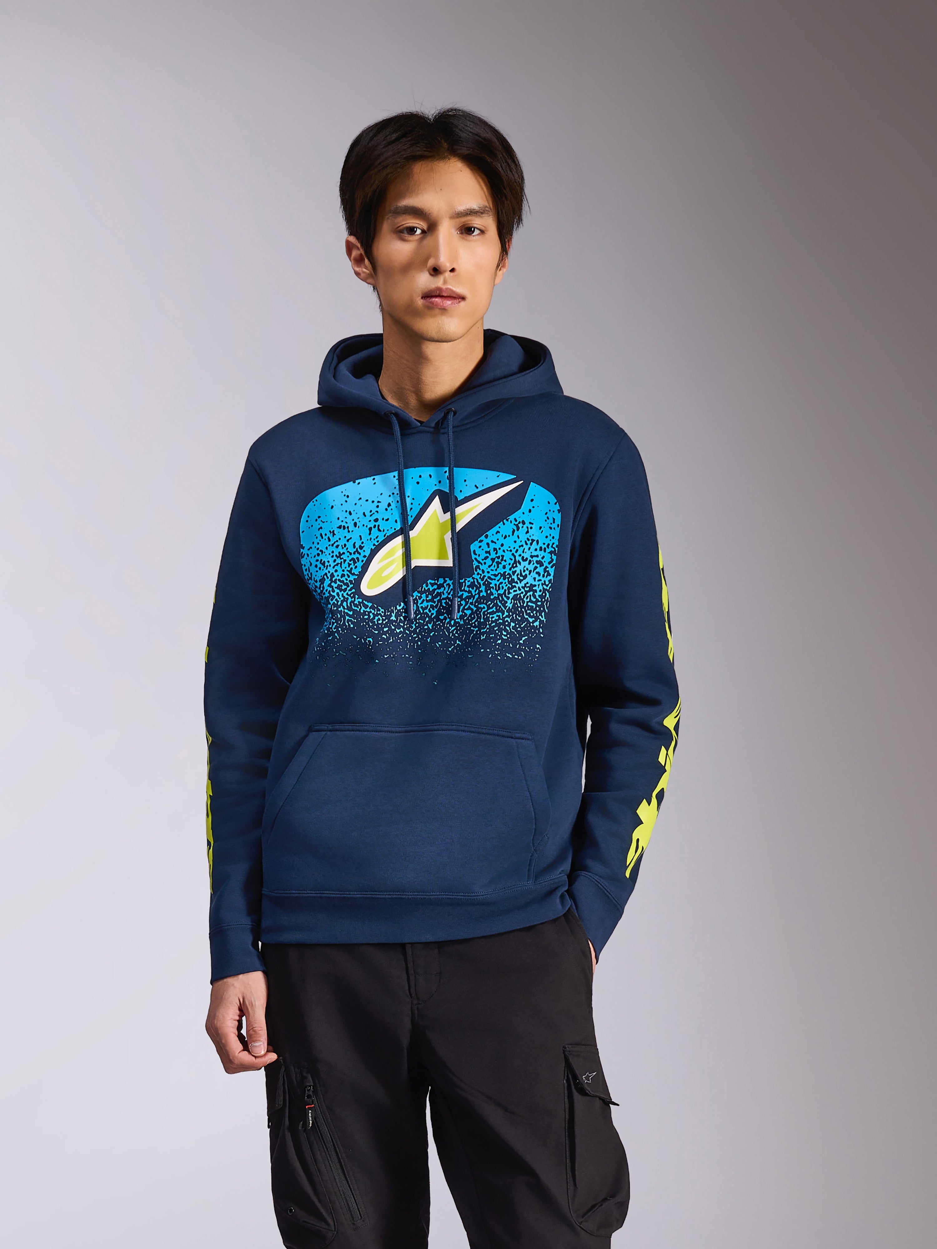 Alpinestars Obsessed Hoodie i marinblått, bärs av en manlig modell, med en limegrön Astars-logotyp med klarblå MX-gradering på bröstet och gula ordlogotyper på ärmarna, avslappnad motorsportinspirerad streetwear.