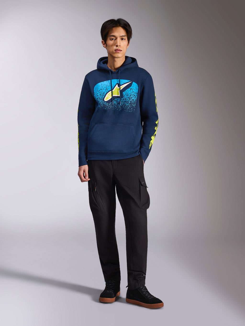 Alpinestars Obsessed Hoodie, marinblå huvtröja, Astars-logga fram med blå MX-gradering och limegul stjärna, limegula Alpinestars-ordloggor på ärmarna, bärs av en modell med svarta cargobyxor och svarta sneakers, avslappnad motorsport-inspirerad streetwear