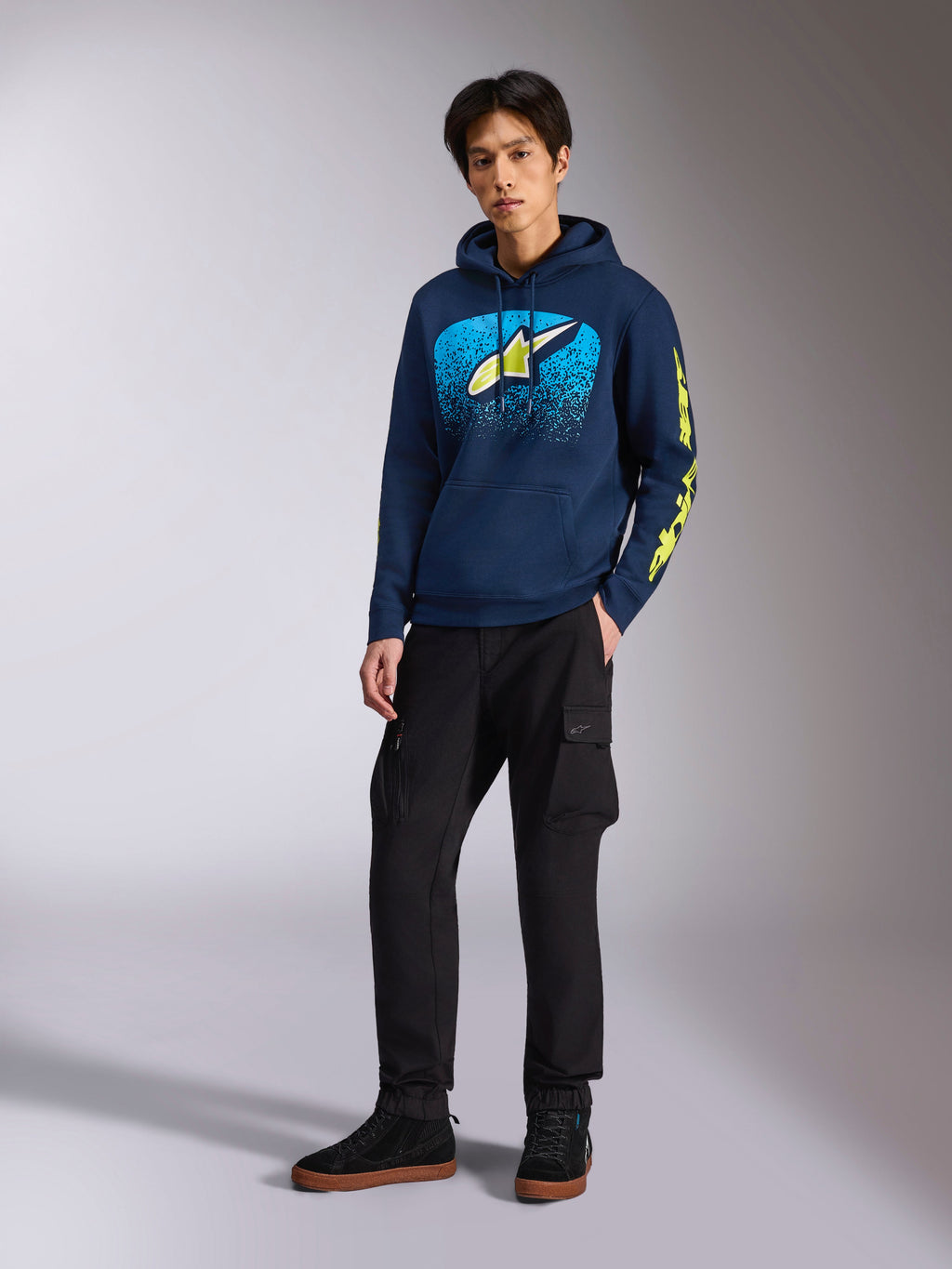 Alpinestars Obsessed Hoodie i marinblått, avslappnad fleecehuvtröja med ljusblå och limegul grafik framtill, limegul Alpinestars-logotyp på ärmen, bärs av en modell med svarta cargobyxor och svarta sneakers, motorsportinspirerat streetwear