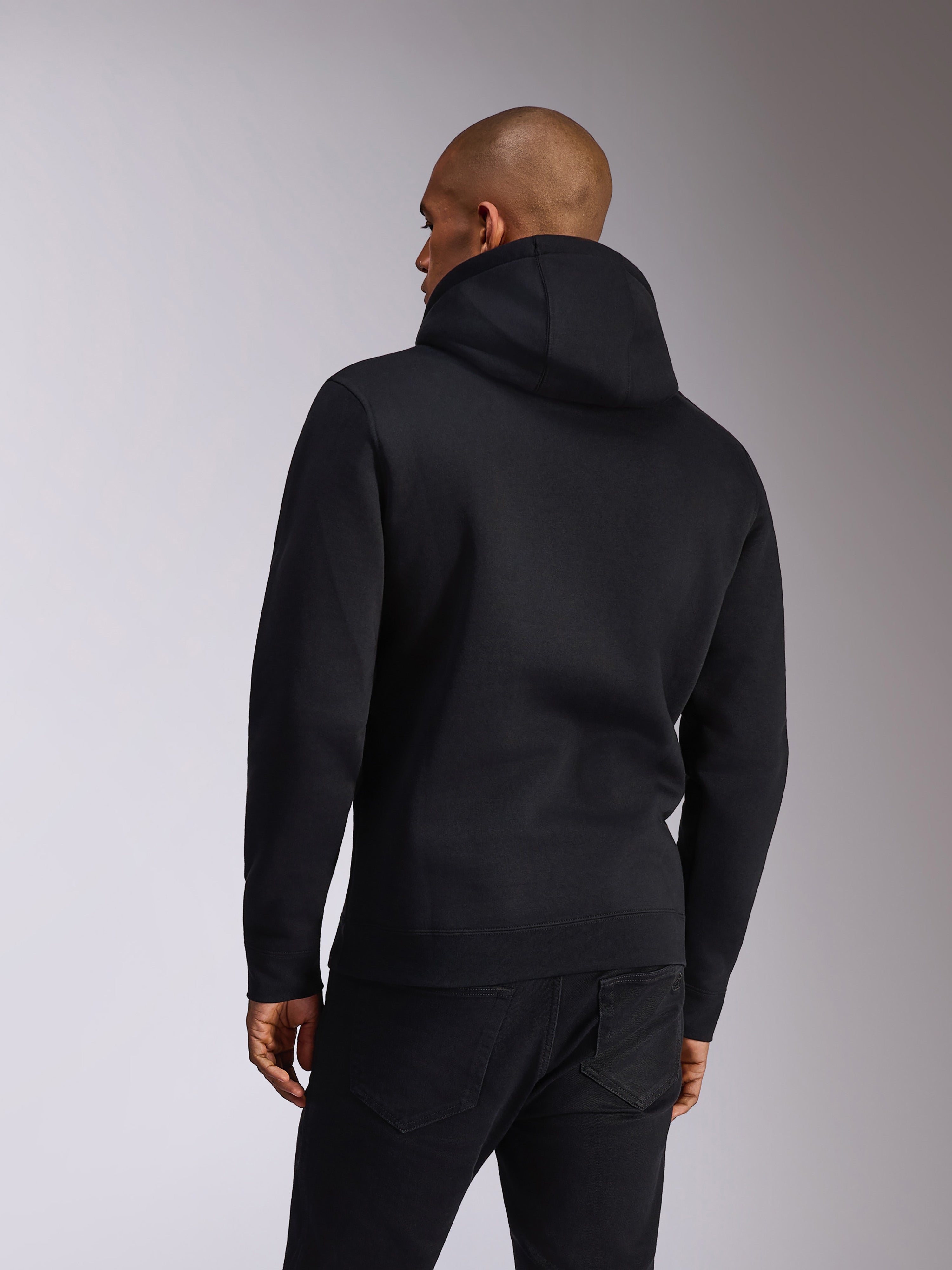 Alpinestars Ellipsoid Hoodie, svart, baksida av en person som bär en huvtröja i fleece, långa ärmar med ribbade muddar och fåll, stylad med svarta jeans, vardagliga motorsportinspirerade sportkläder