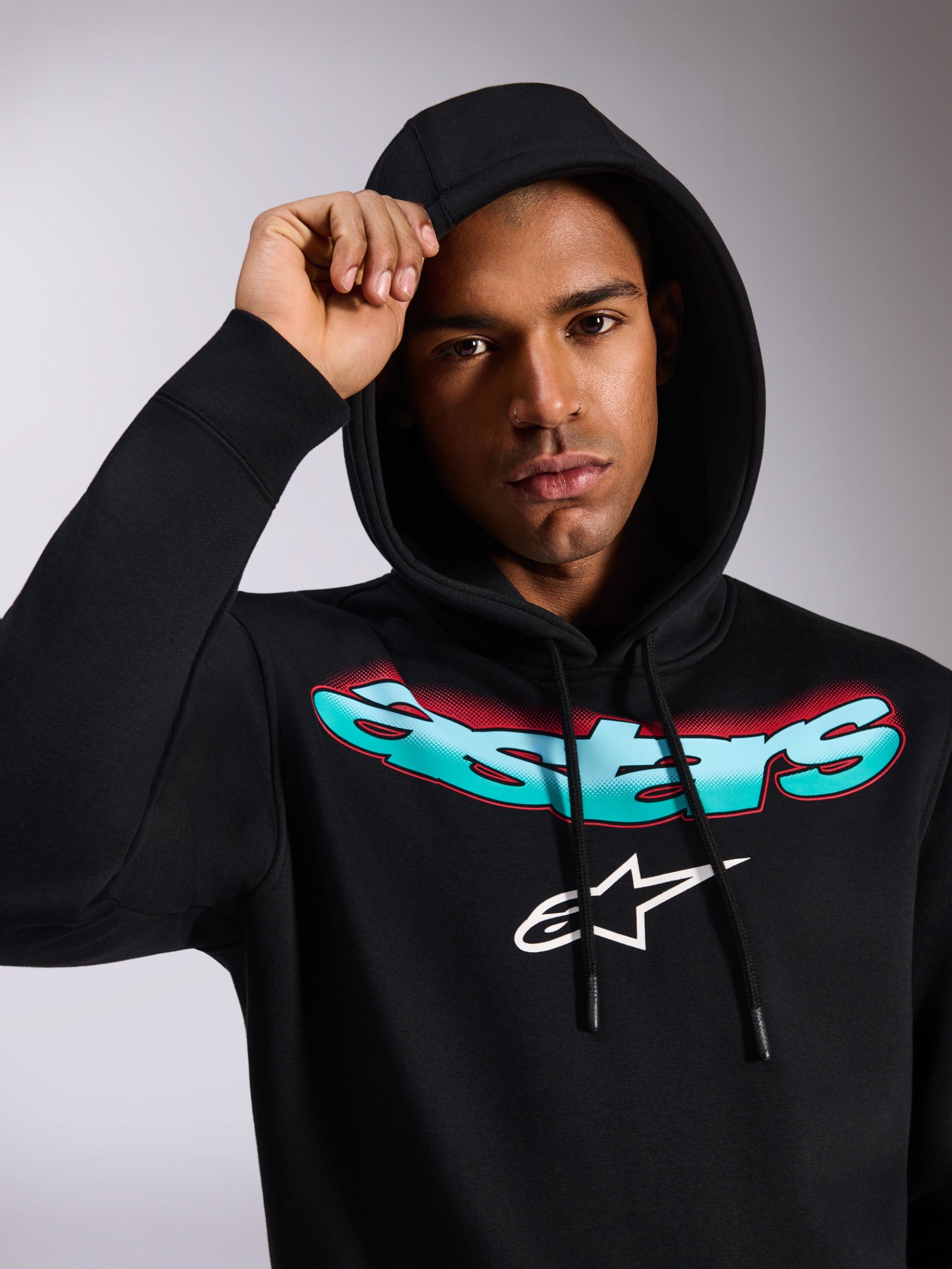 Alpinestars Ellipsoid Hoodie, svart huvtröja i fleece, med stiliserad Astars-text i turkos och röd färggradient och vit logotyp på bröstet, bärs av modell med huvan uppfälld, avslappnad motorsportinspirerad streetwear