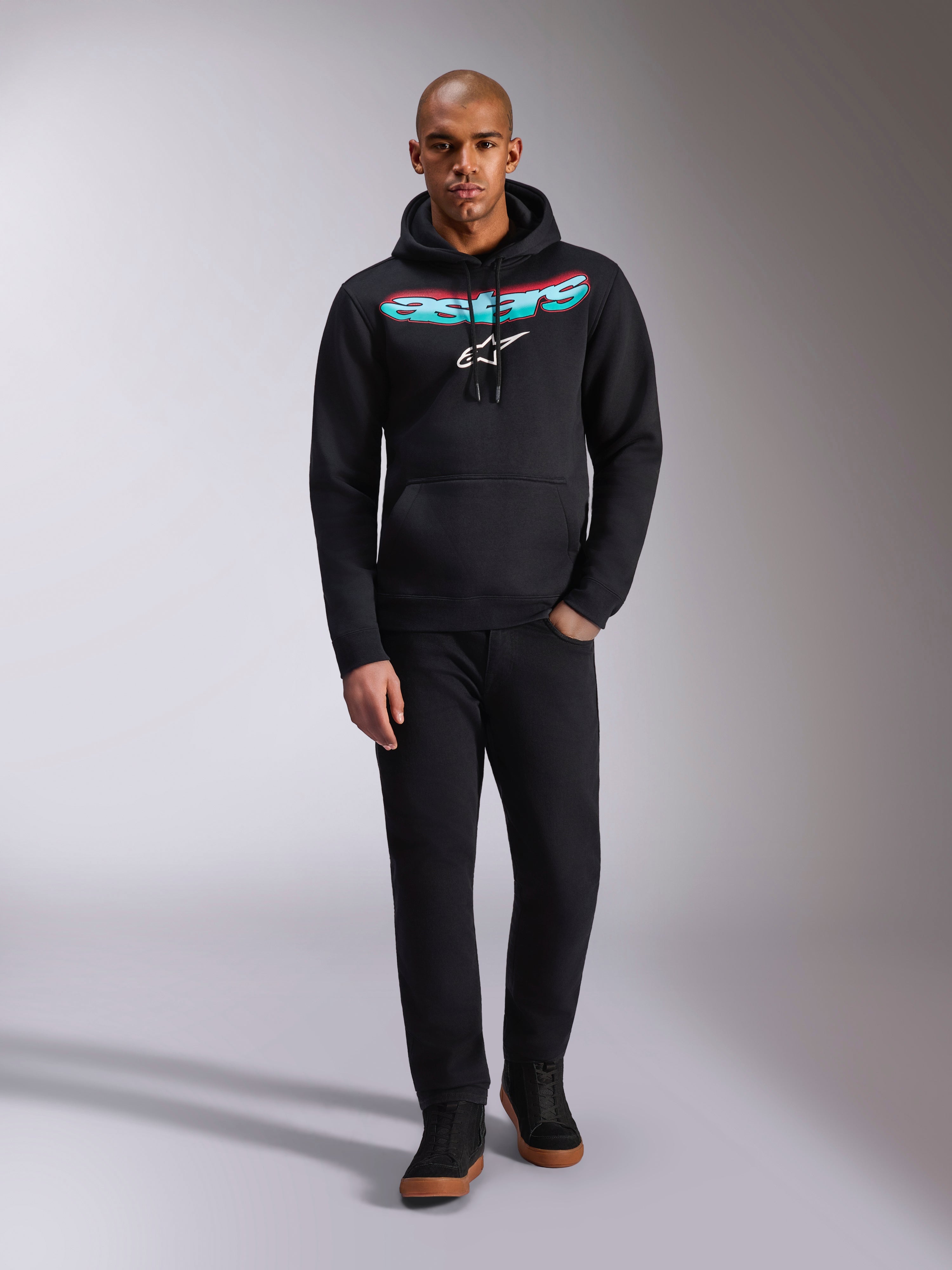 Alpinestars Ellipsoid Hoodie, svart huvtröja i fleece med en böjd Astars-text i en röd och blågrön färggradient och vit stjärnlogotyp, bärs av modell med svarta byxor och sneakers, casual motorsport-streetwear