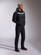 Alpinestars Ellipsoid Hoodie, svart huvtröja i fleece med en böjd blågrön och röd tonad Astars-text och vit stjärnlogotyp, bärs av en modell med svarta byxor, avslappnad motorsportinspirerad streetwear