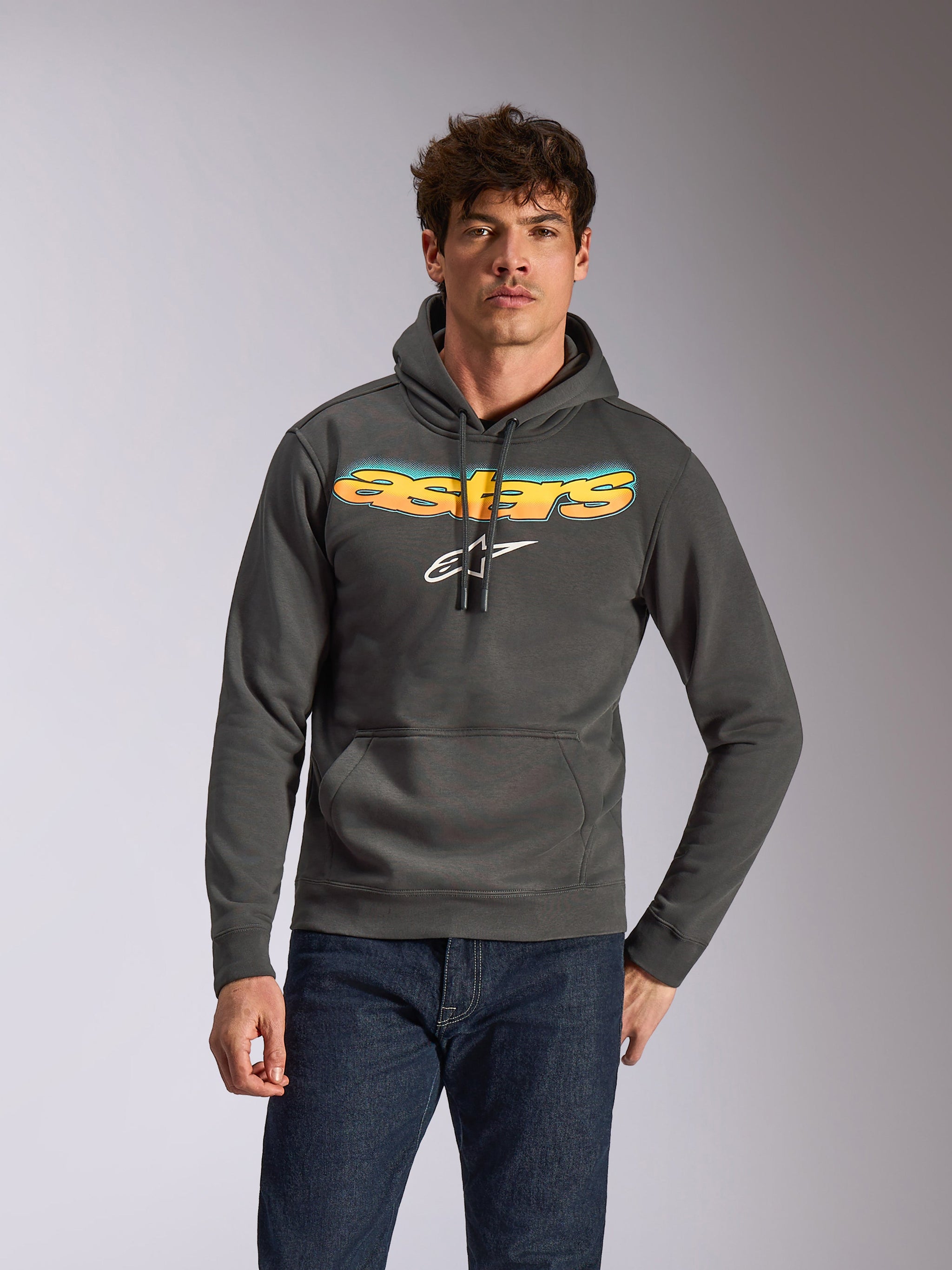 Alpinestars Ellipsoid Hoodie, avslappnade sportkläder, gunmetal-grå med gul och blå färggradient på Astars-texten och vit logotyp, pullover-modell med huva och dragsnöre, bärs med mörkblå jeans