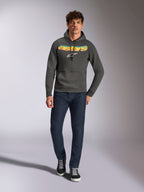 Alpinestars Ellipsoid Hoodie, avslappnade sportkläder, gunmetal-grå med orange och gul färggradient på Astars-logotypen och vit symbol, huva med dragsnöre, bärs av en manlig modell med mörkblå jeans och svarta sneakers, modernt motorsportinspirerat streetwear