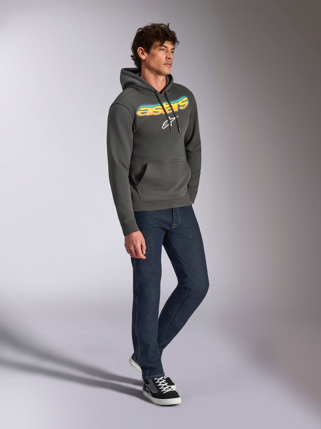 Alpinestars Ellipsoid Hoodie, gunmetal-grå huvtröja i fleece, böjd Astars-text på framsidan med blå och gul färggradient och vit logotyp, stylad med mörka jeans och svarta sneakers, vardaglig streetwear.