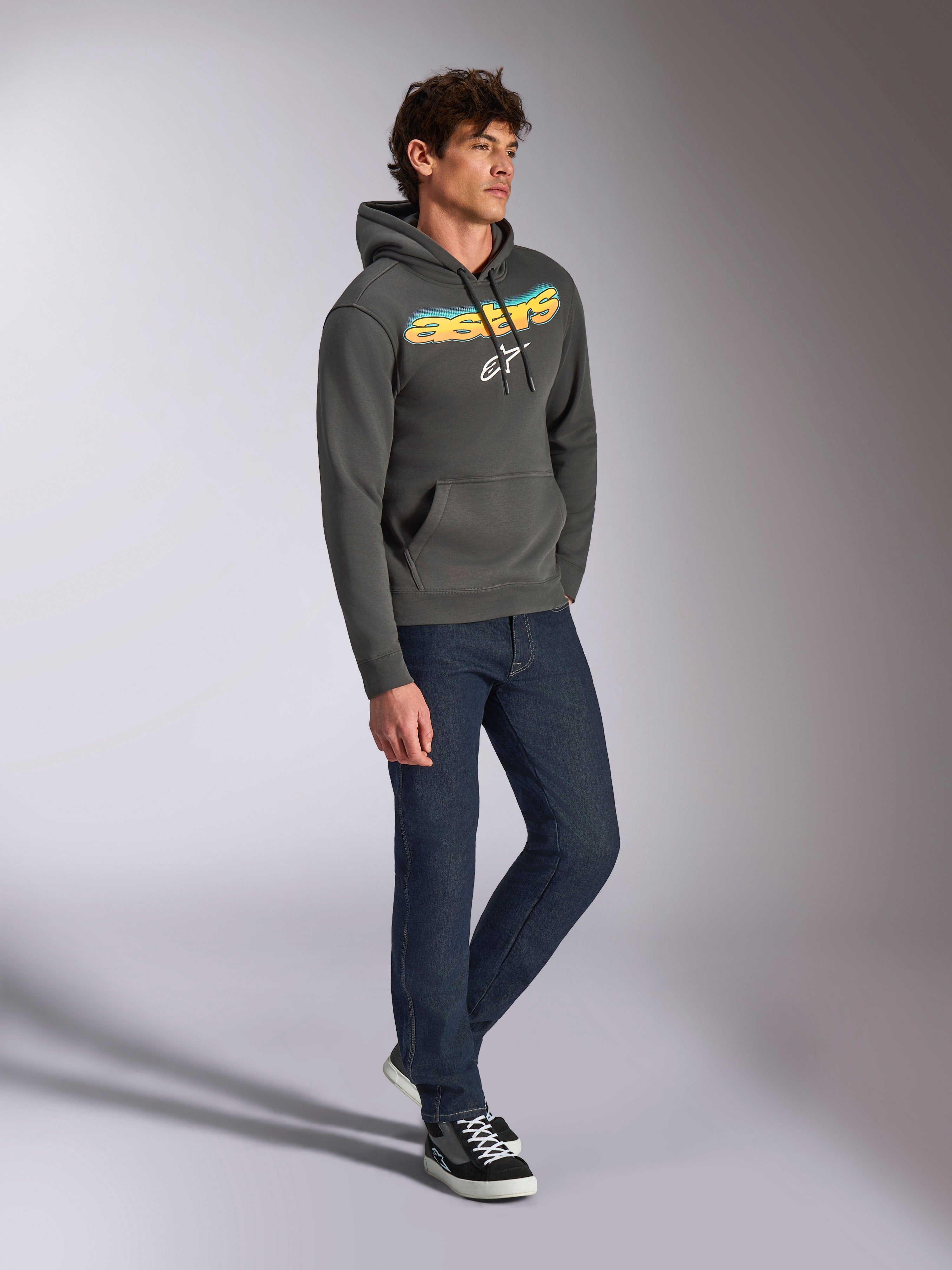 Alpinestars Ellipsoid Hoodie, gunmetal-grå huvtröja i fleece, böjd Astars-text på framsidan med blå och gul färggradient och vit logotyp, stylad med mörka jeans och svarta sneakers, vardaglig streetwear.
