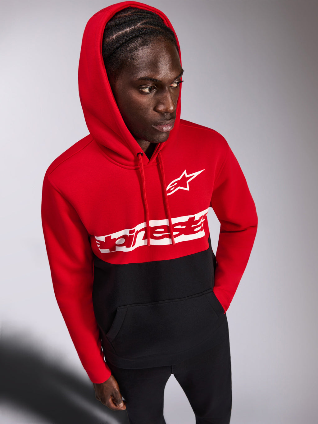 Alpinestars Parallel Hoodie, vardaglig huvtröja i fleece, röd och svart färgblockerad design, vit Alpinestars ordlogotyp och Astars-logotyp på bröstet, bärs av en man med huvan uppe och svarta byxor, motorsport-inspirerat streetwear
