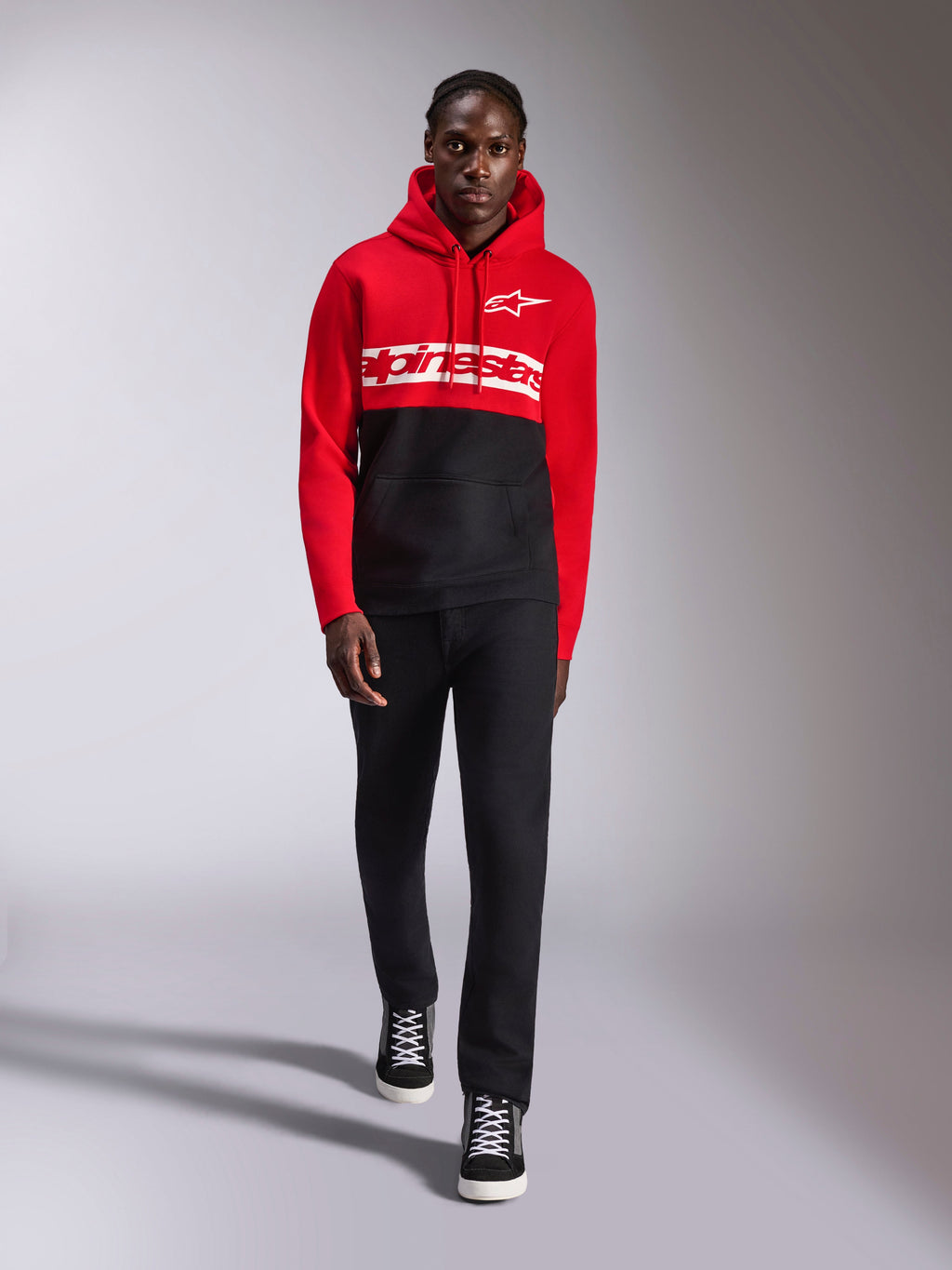 Alpinestars Parallel Huvtröja, färgblockad huvtröja i fleece, röd och svart, med vit Alpinestars-ordlogga över bröstet och Astars-logga på vänster bröst, bärs av en modell med svarta byxor och high-top sneakers, avslappnad motorsport-streetwear