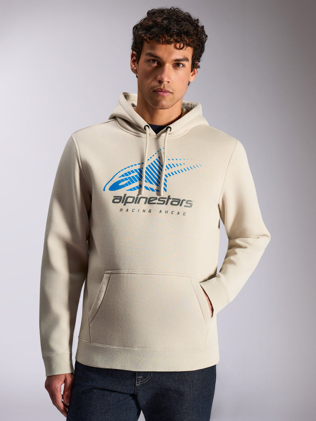 Alpinestars Pulse huvtröja, casual streetwear, sandfärgad, blå och grå Astars Combo-logotyp på framsidan med speed line fade och texten 'Racing Ahead', huva med dragsko, känguruficka, bärs av en modell med mörka jeans