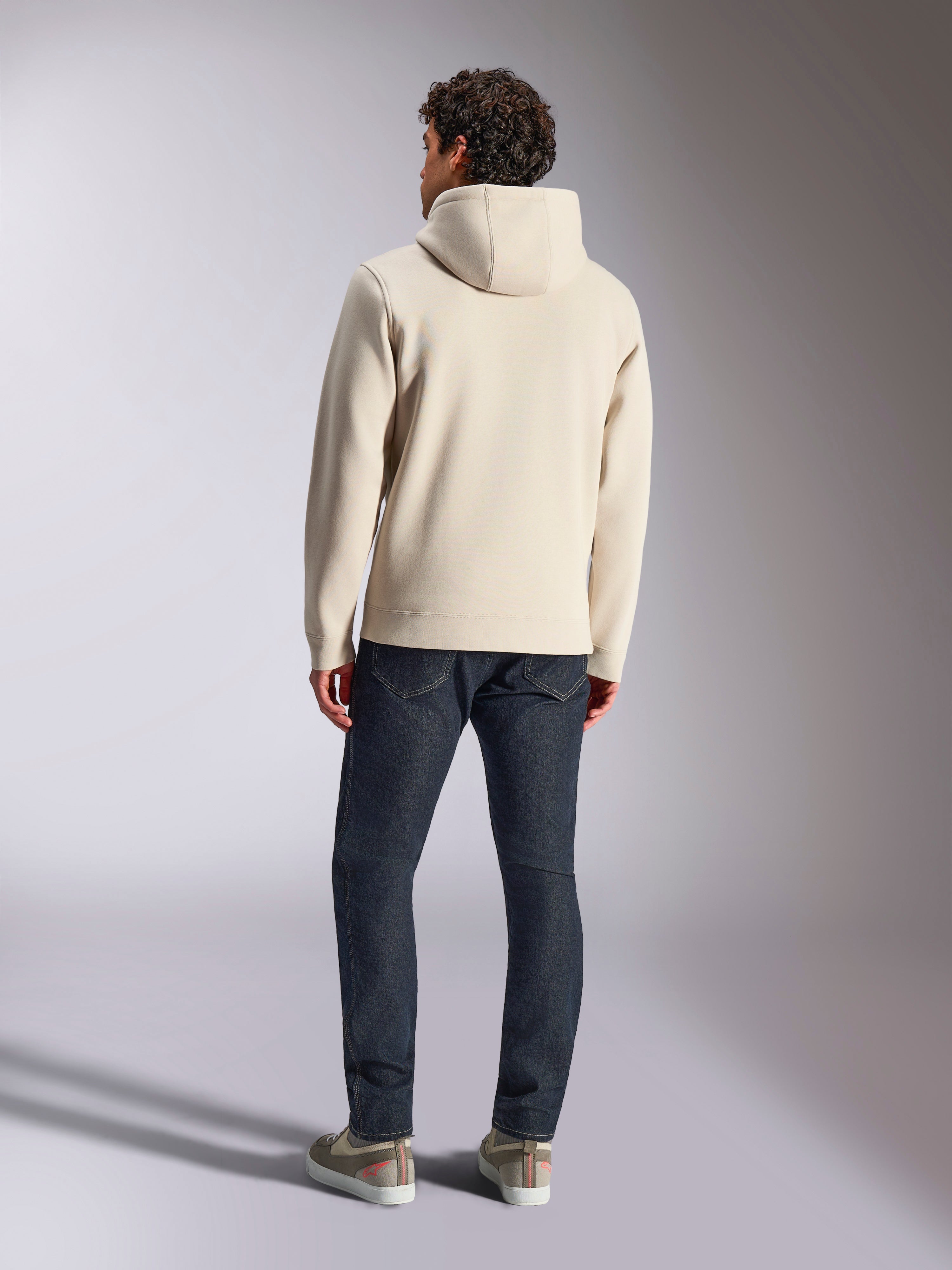 Alpinestars Pulse Hoodie, Off White, baksida av en man som bär en avslappnad pullover-huvtröja i fleece, mörka jeans och sneakers, modern sportklädesdesign