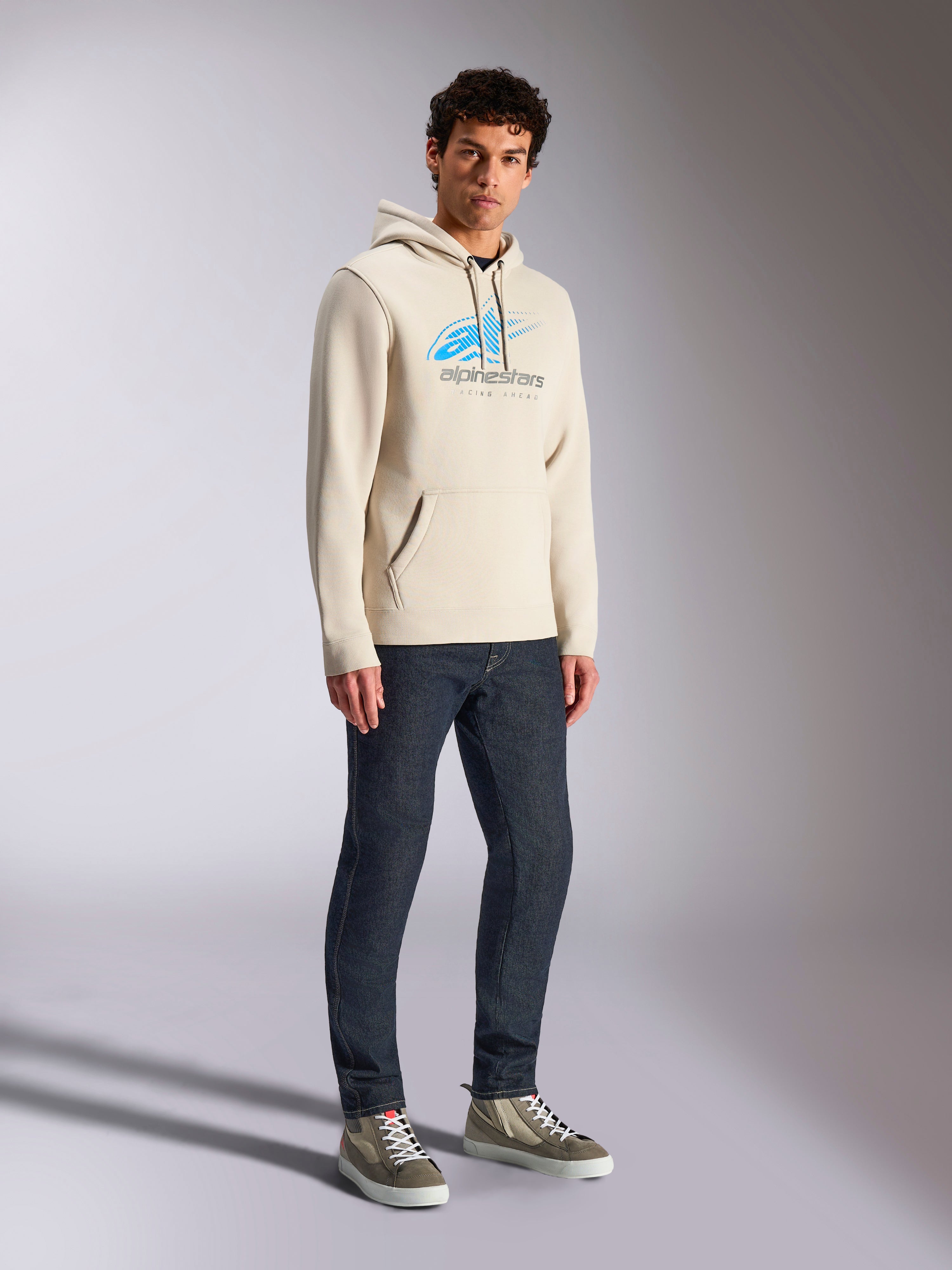 Alpinestars Pulse Hoodie, Off White casual streetwear, huvtröja i fleece med en blå och grå Astars-logotyp med speed line fade, känguruficka, bärs av modell med mörka denimjeans och high-top sneakers.