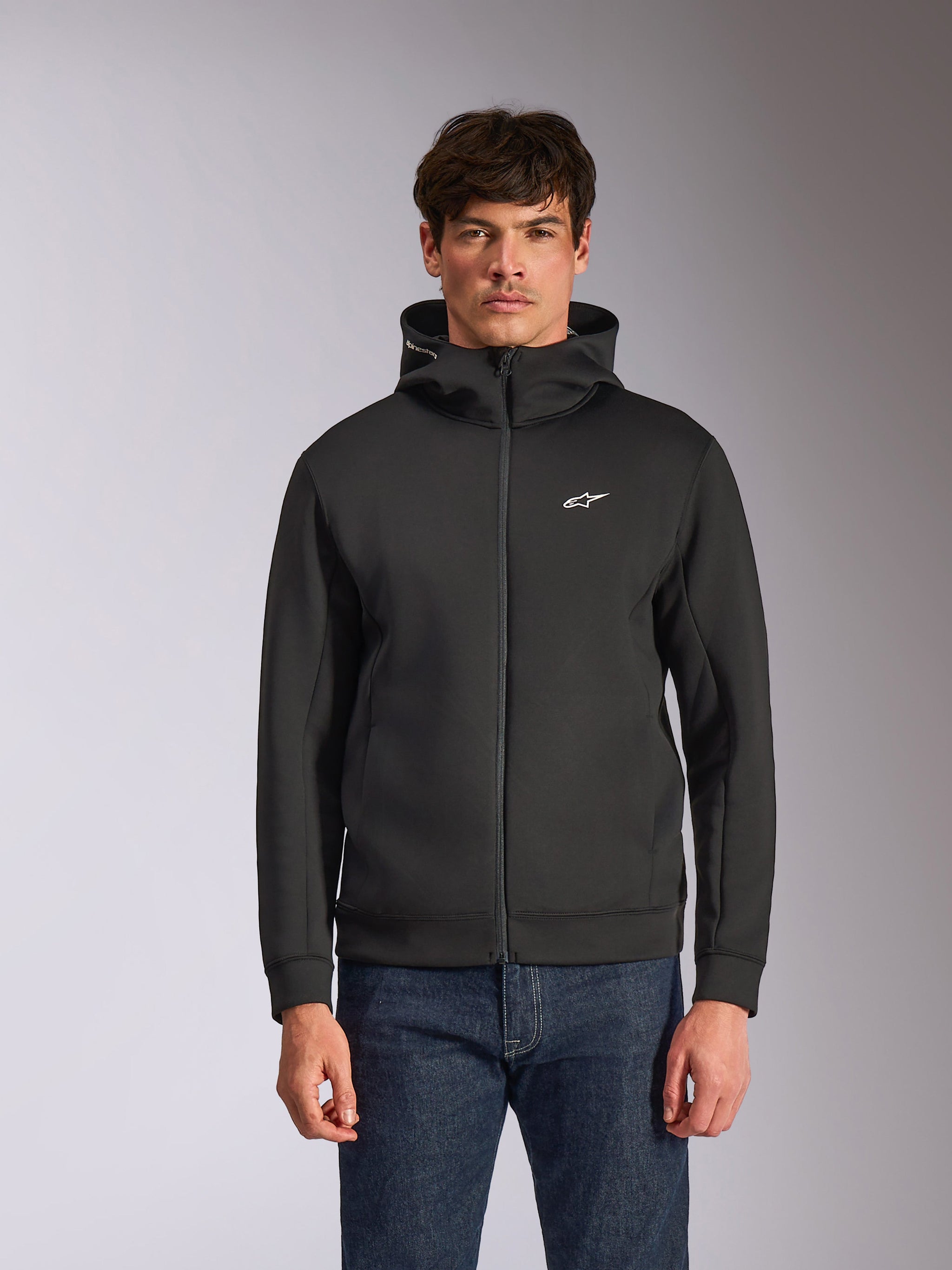 Alpinestars Cypher Hoodie, svart fleecehuvtröja med dragkedja fram, med en huva med hög krage, vit branding och en liten vit Astars-logotyp på bröstet, bärs av en modell i blå jeans, avslappnad motorsport-inspirerad streetwear.