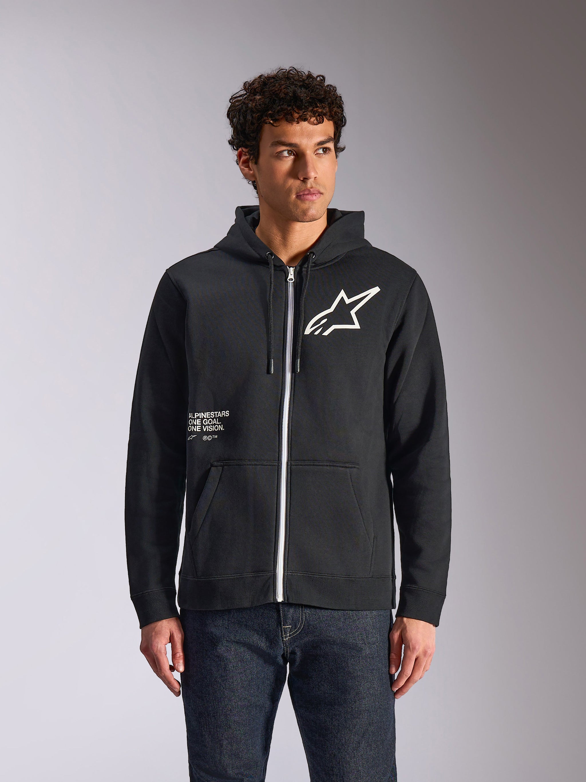 Alpinestars Transverse Hoodie, svart huvtröja i fleece med dragkedja fram, vit Astars-logotyp och One Goal-grafik på framsidan, bärs av en modell med mörka jeans, vardaglig motorsport-inspirerad streetwear