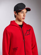 Alpinestars Transverse Hoodie, röd fleecehuvtröja med dragkedja fram, svart Astars-logotyp på bröstet och texten 'One Goal. One Vision.' nedtill på framsidan, casual motorsport-streetwear, bärs av en modell med en svart keps