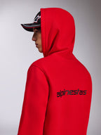 Alpinestars Transverse Hoodie, röd huvtröja i fleece, vy snett bakifrån som visar den svarta Alpinestars-ordlogotypen på ryggen, bärs med huvan uppe och en svart keps, avslappnad motorsportinspirerad streetwear