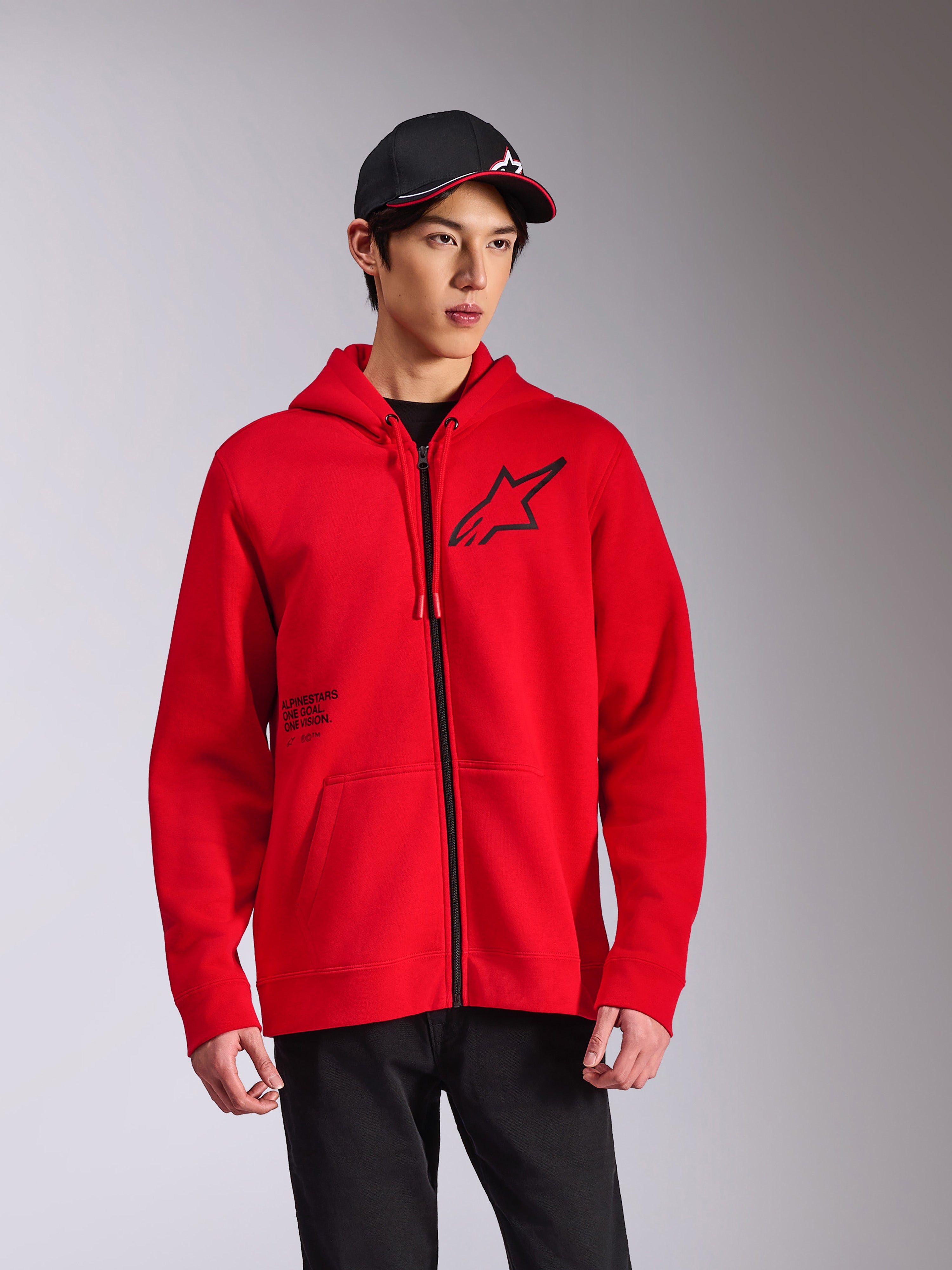 Alpinestars Transverse Hoodie, röd fleecehuvtröja med dragkedja fram, svart Astars-logotyp på bröstet och 'One Goal'-tryck, avslappnad streetwear-stil buren med en svart keps och byxor