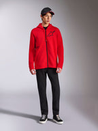 Alpinestars Transverse Hoodie, röd fleecehuvtröja med hel dragkedja, svart Astars-logotyp på bröstet och One Goal-tryck på sidan, bärs av manlig modell med svarta byxor och keps, avslappnad motorsport-inspirerad streetwear