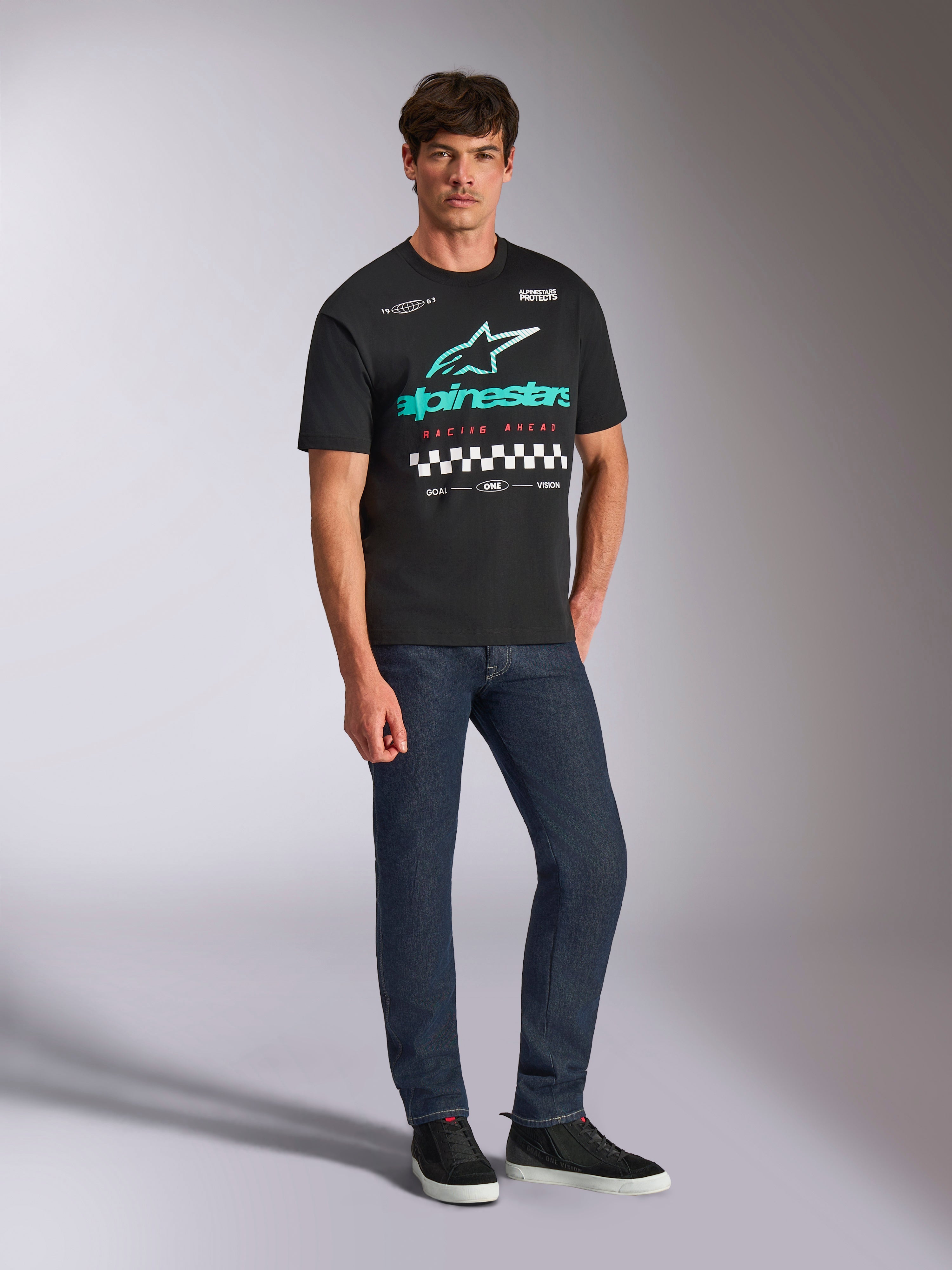 Alpinestars Aplomb Oversized CSF Tee, kortärmad t-shirt, svart, stor turkos och vit Alpinestars-logotyp med färggradient, Racing Ahead-text och schackrutigt mönster, bärs med mörkblå jeans och svarta sneakers, avslappnad motorsport-streetwear