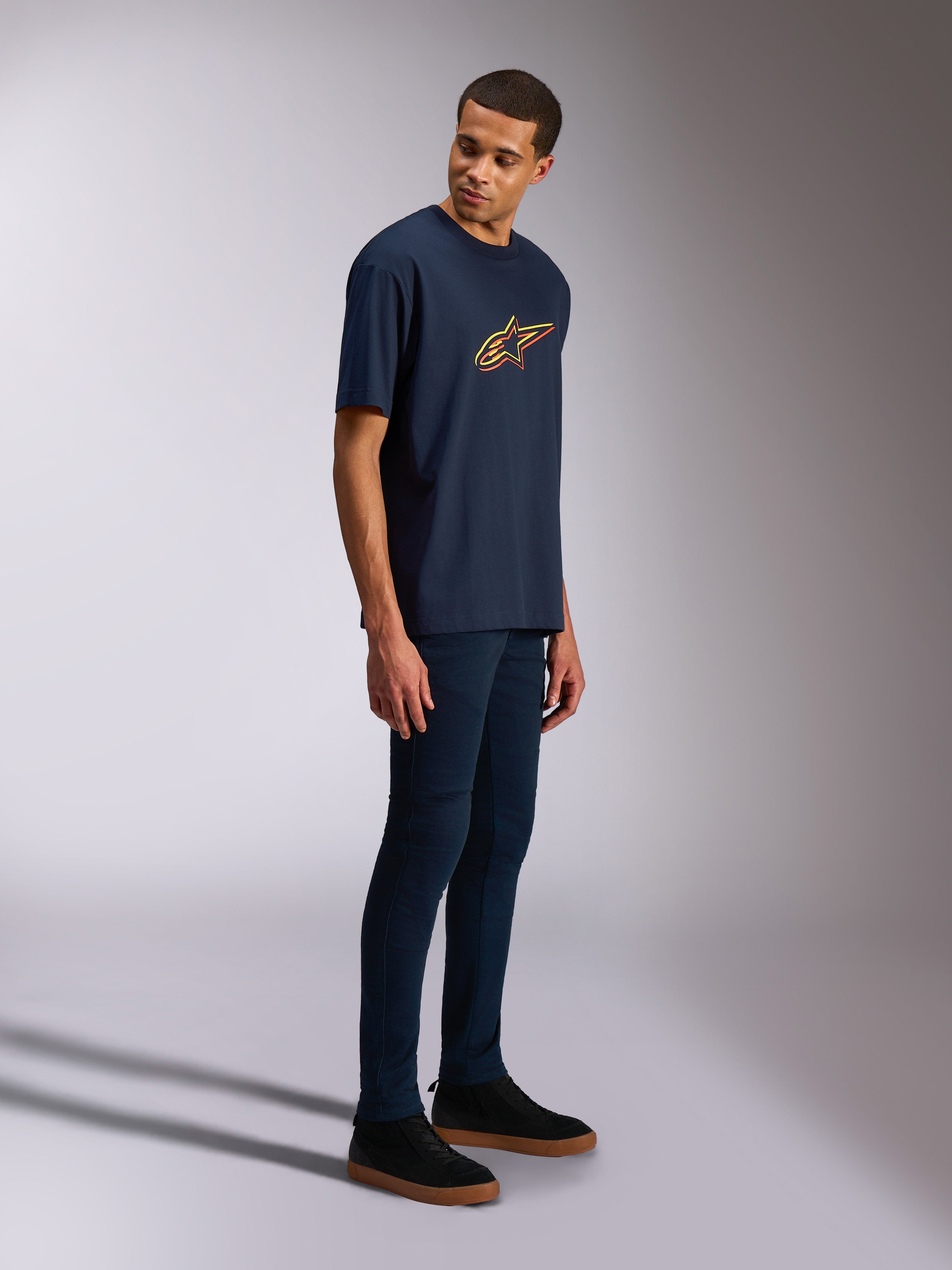 Alpinestars Alter Oversized CSF T-shirt, kortärmad t-shirt, marinblå, gul och orange Astars-logotyp, bärs av en modell med mörka jeans och svarta sneakers, avslappnad motorsportinspirerad streetwear