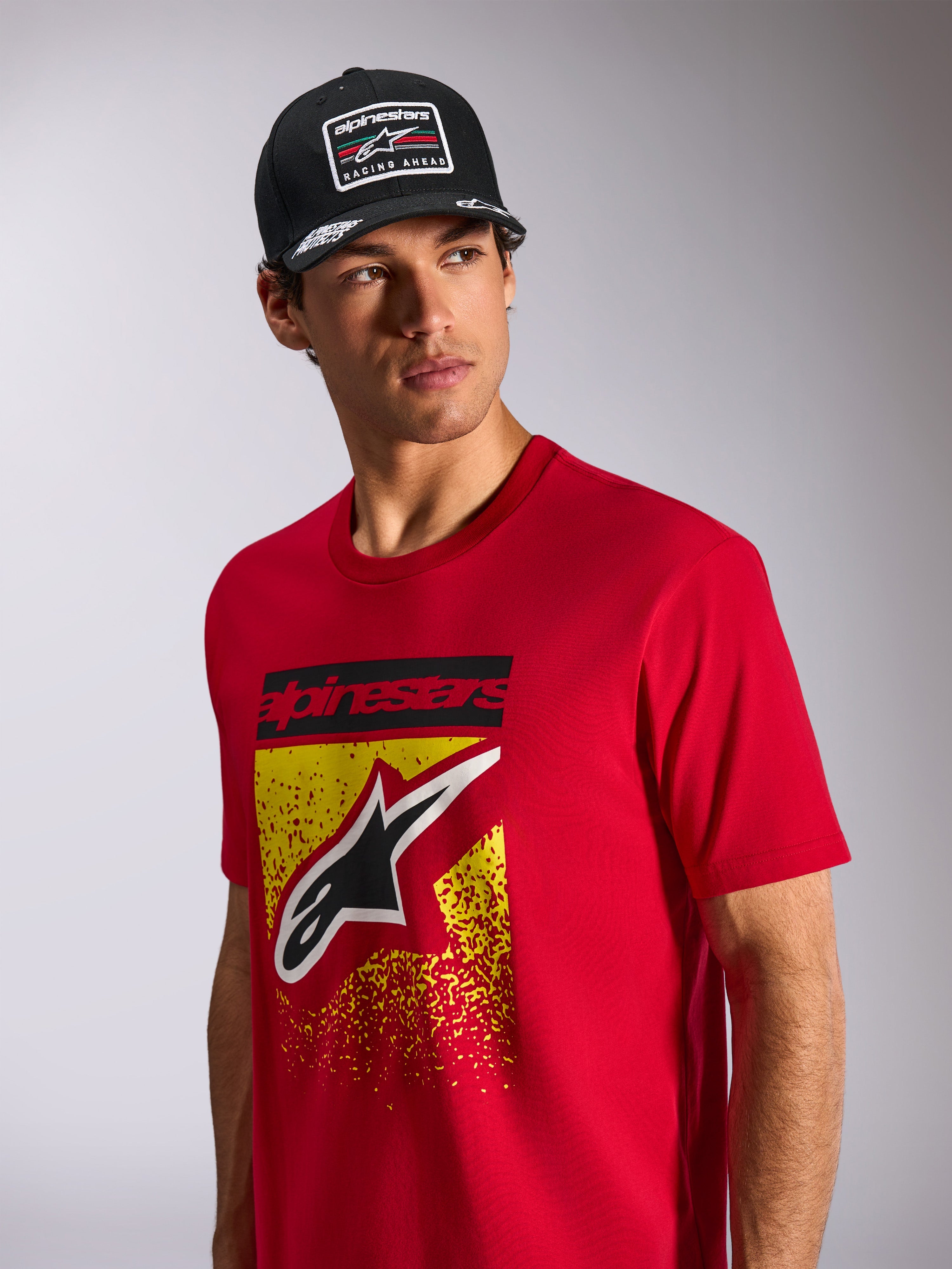 Alpinestars Noka CSF Tee, kortärmad t-shirt, röd, med Astars-logotyp på framsidan med gul och röd MX-gradering, bärs med en svart Alpinestars 'Racing Ahead'-keps, avslappnad motorsport-streetwear