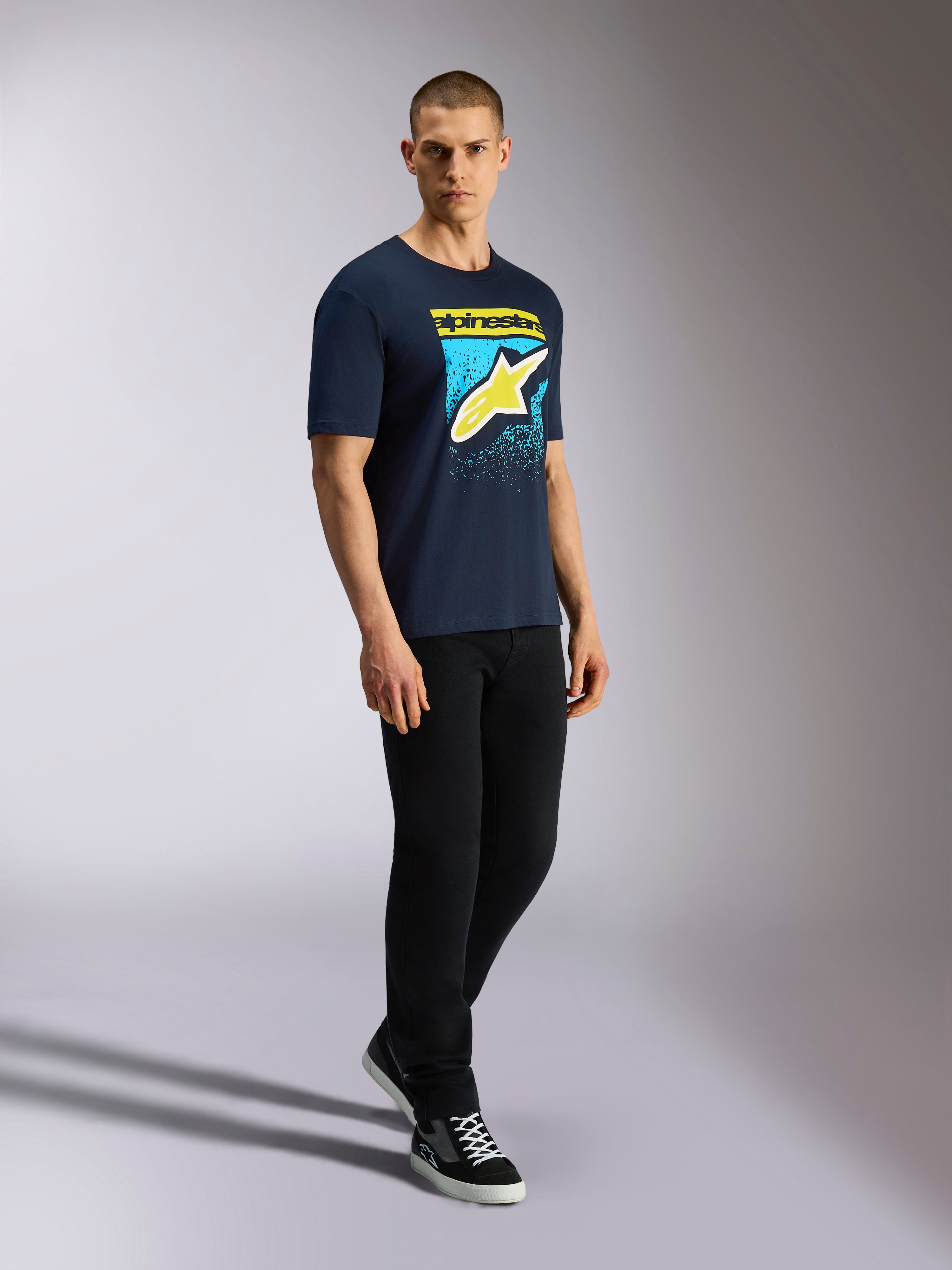 Alpinestars Noka CSF Tee, kortärmad t-shirt, marinblå, gul Astars-logotyp med blå MX-graderad grafik, manlig modell i marinblå t-shirt, svarta byxor och sneakers, avslappnad motorsport-streetwear