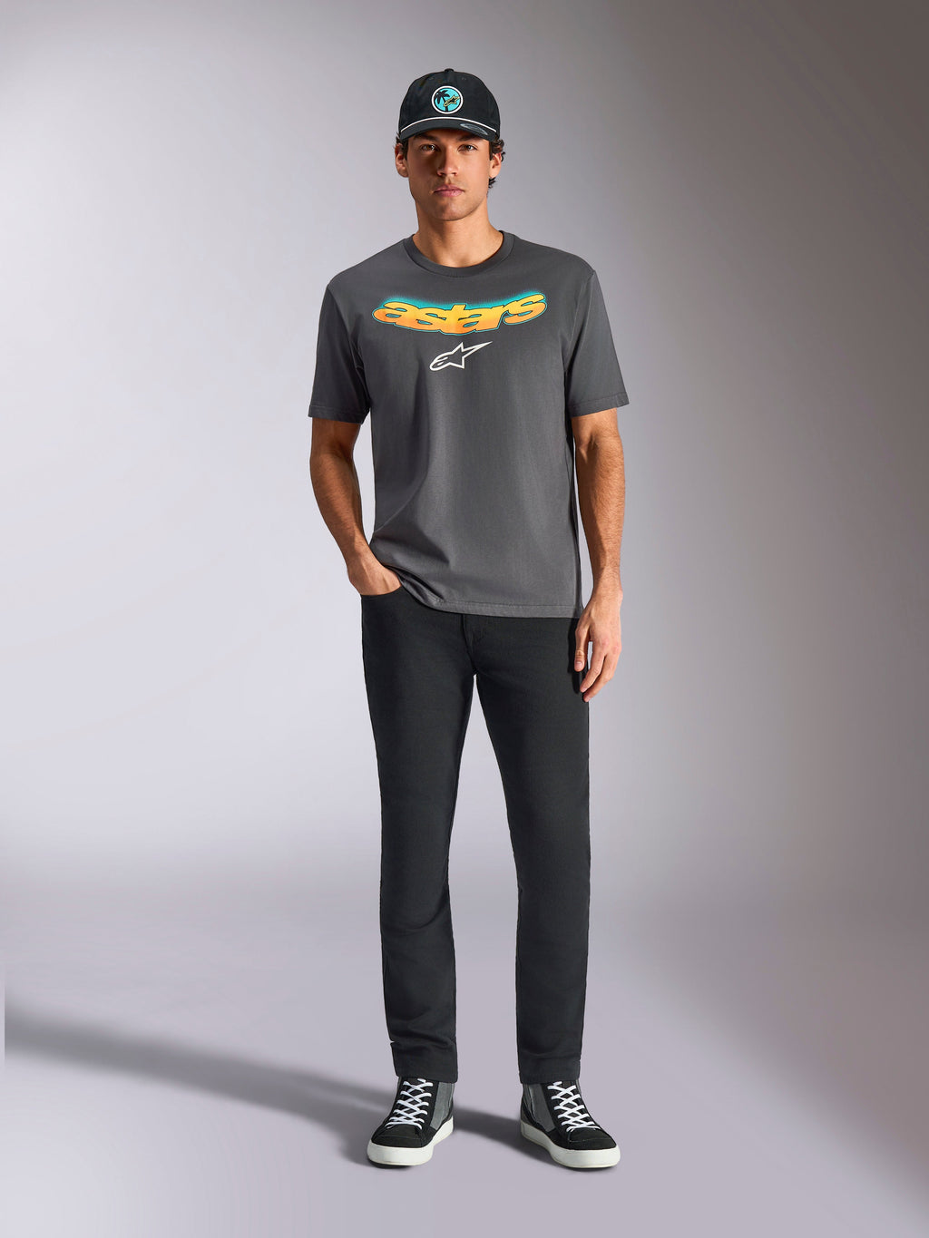 Alpinestars Ellipsoid CSF Tee, kortärmad t-shirt, mörkgrå, med en böjd gul och orange gradient-text och vit Astars-logotyp, bärs av en modell med svarta byxor och en baseballkeps, casual sportswear-stil