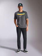 Alpinestars Ellipsoid CSF Tee, kortärmad t-shirt, mörkgrå, med en böjd gul och orange gradient-text och vit Astars-logotyp, bärs av en modell med svarta byxor och en baseballkeps, casual sportswear-stil