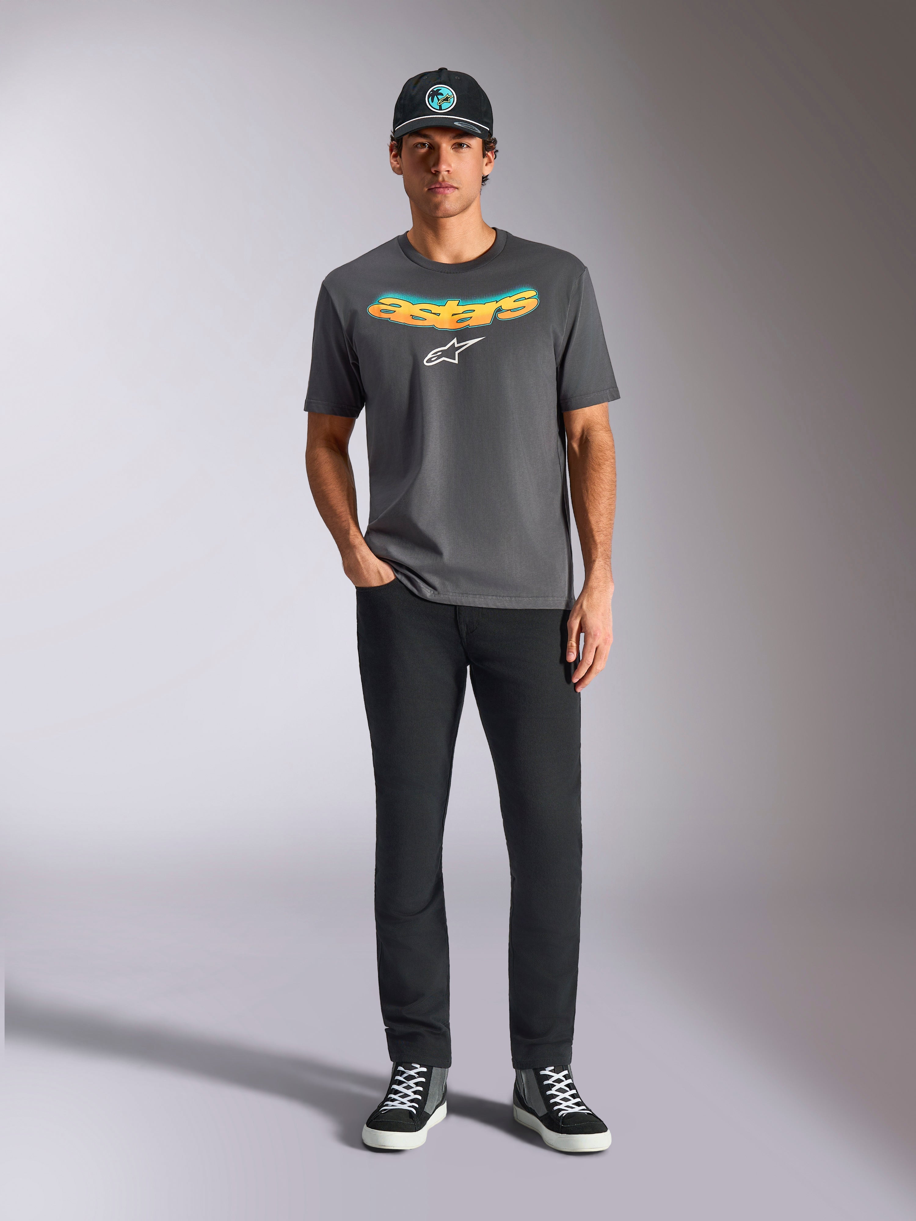 Alpinestars Ellipsoid CSF Tee, kortärmad t-shirt, mörkgrå, med en böjd gul och orange gradient-text och vit Astars-logotyp, bärs av en modell med svarta byxor och en baseballkeps, casual sportswear-stil