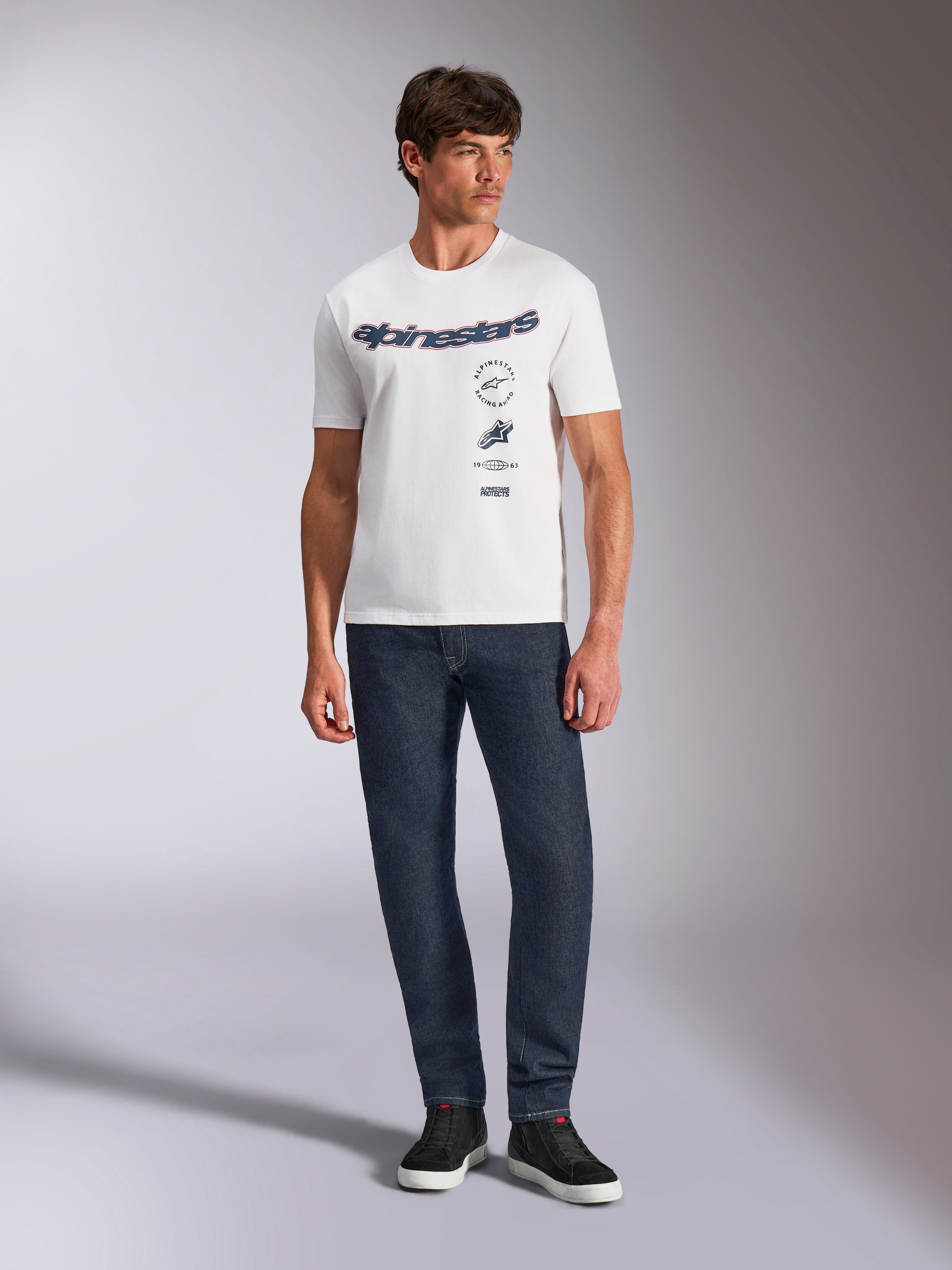 Alpinestars Abus CSF Tee, Short Sleeve T-Shirt, White, featuring curved Alpinestars logo and multi-stacked graphics on the chest, worn by a male model with dark blue jeans and black sneakers, casual motorsport streetwear