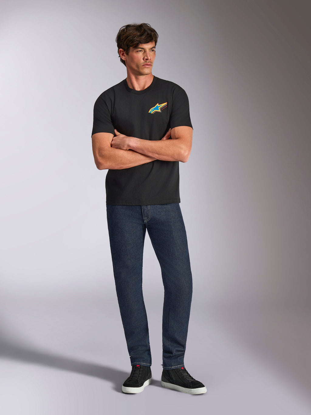 Alpinestars Angler CSF Tee, Short Sleeve T-Shirt, Black, Vibrant Multi-Color Astars Logo on Chest, worn by model with arms crossed, dark denim jeans and black sneakers, casual motorsport streetwear