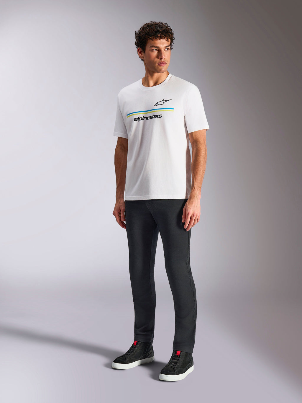 Alpinestars Lanes CSF Tee, vit kortärmad t-shirt, bärs av modell med svarta slim-fit byxor och svarta high-top sneakers, med blå och gula horisontella ränder och Alpinestars-logotyp på bröstet, avslappnad motorsport-inspirerad streetwear