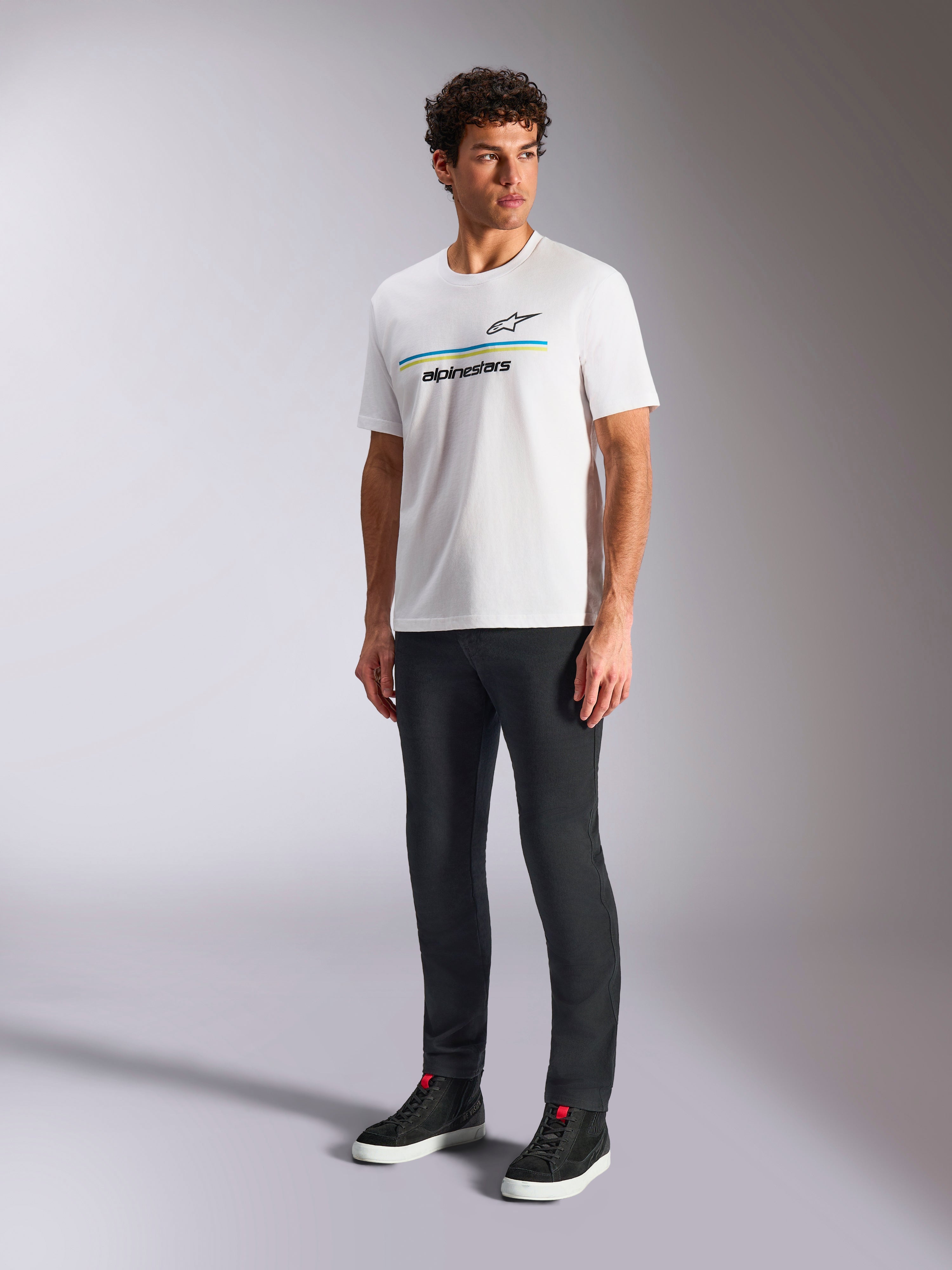Alpinestars Lanes CSF Tee, vit kortärmad t-shirt, bärs av modell med svarta slim-fit byxor och svarta high-top sneakers, med blå och gula horisontella ränder och Alpinestars-logotyp på bröstet, avslappnad motorsport-inspirerad streetwear