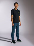 Alpinestars Order CSF Tee, kortärmad t-shirt, svart, med gröna hjälm- och palmträdslogotyper på bröstet, avslappnad motorsport-streetwear, bärs med blå jeans och svarta sneakers
