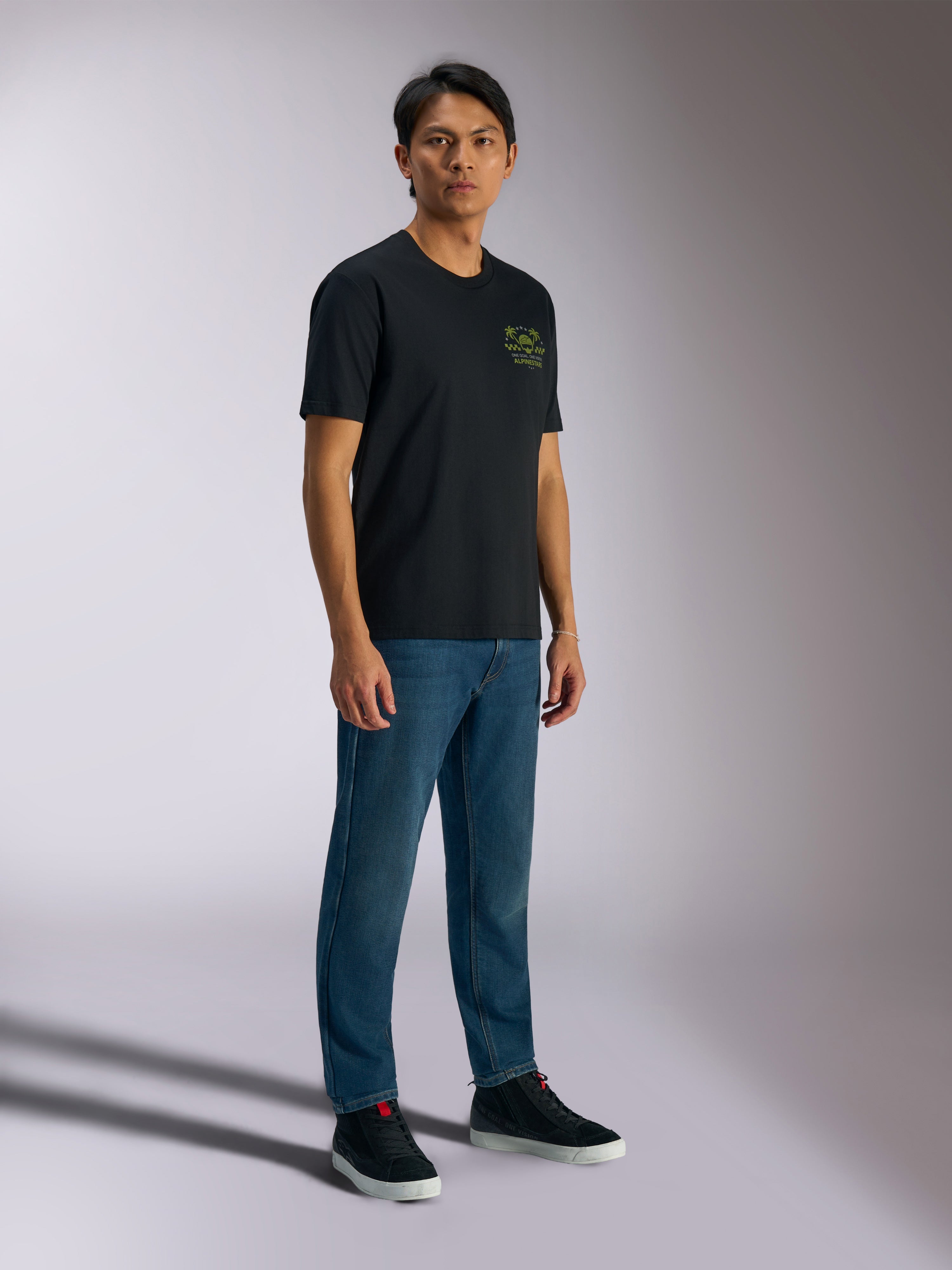Alpinestars Order CSF Tee, kortärmad t-shirt, svart, med gröna hjälm- och palmträdslogotyper på bröstet, avslappnad motorsport-streetwear, bärs med blå jeans och svarta sneakers