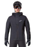 Alpinestars A-Dura Elite vattentät jacka, svart MTB-jacka, design med hög krage och huva, dragkedja fram med skyddande bröstflik och vit logotyp, bärs med svart hjälm och cykelhandskar, tekniskt väderskydd för trailcykling