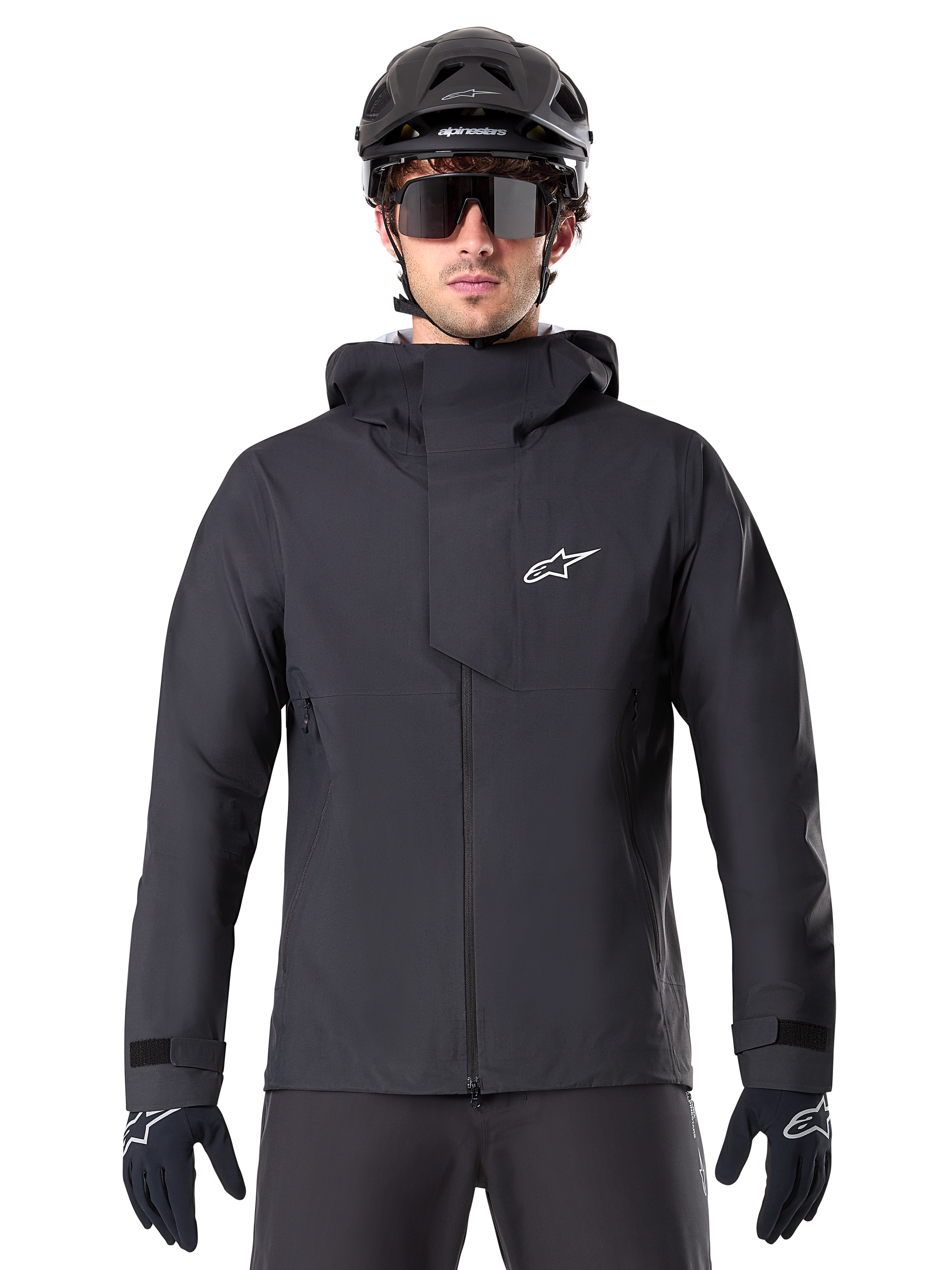 Alpinestars A-Dura Elite vattentät jacka, svart MTB-jacka, design med hög krage och huva, dragkedja fram med skyddande bröstflik och vit logotyp, bärs med svart hjälm och cykelhandskar, tekniskt väderskydd för trailcykling