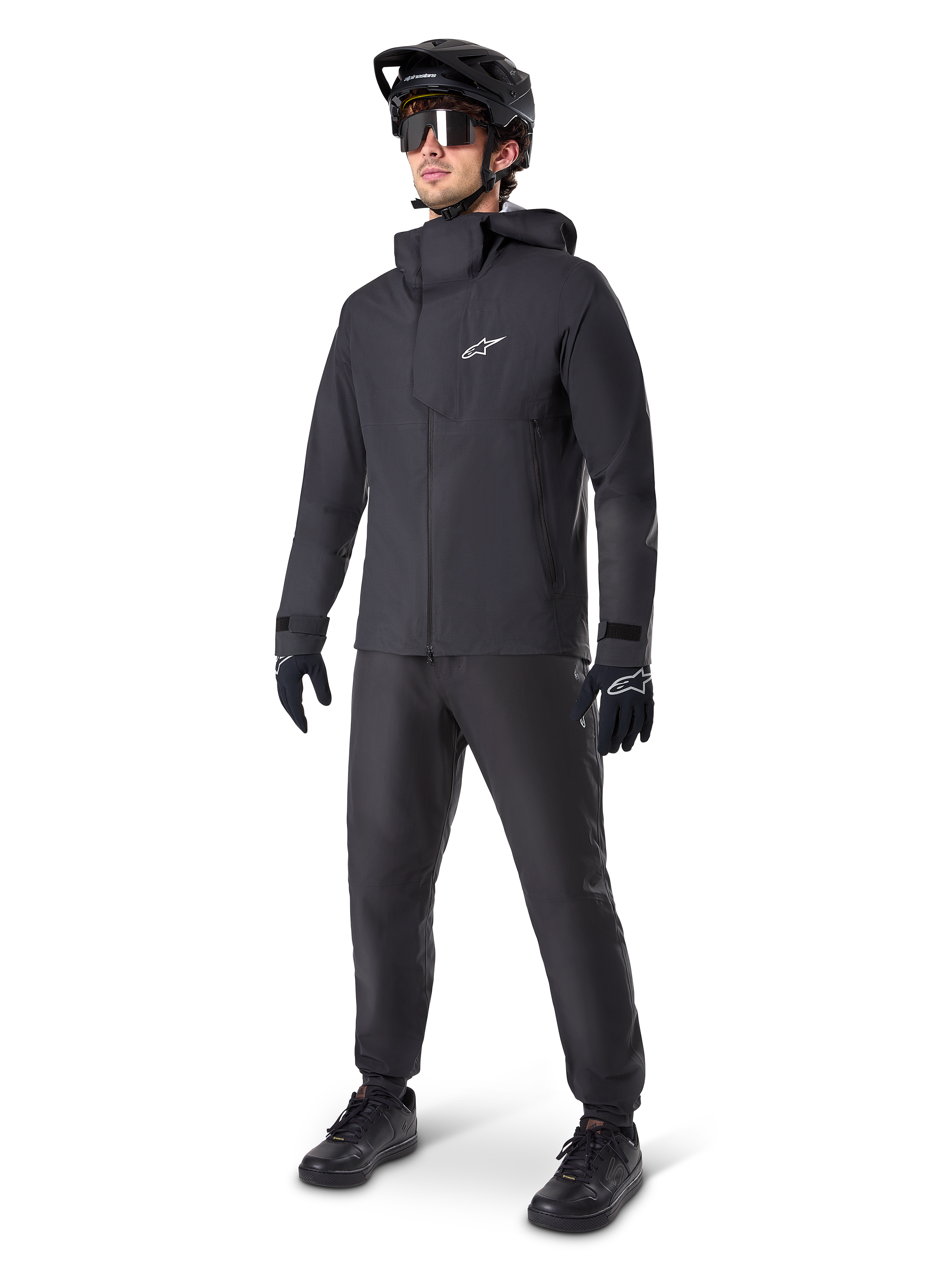Alpinestars A-Dura Elite vattentät jacka i svart, bärs av en manlig cyklist med matchande svarta MTB-byxor, handskar och hjälm. Jackan har en teknisk vattentät design med en vit Astars-logotyp på bröstet och en hög skyddande krage.