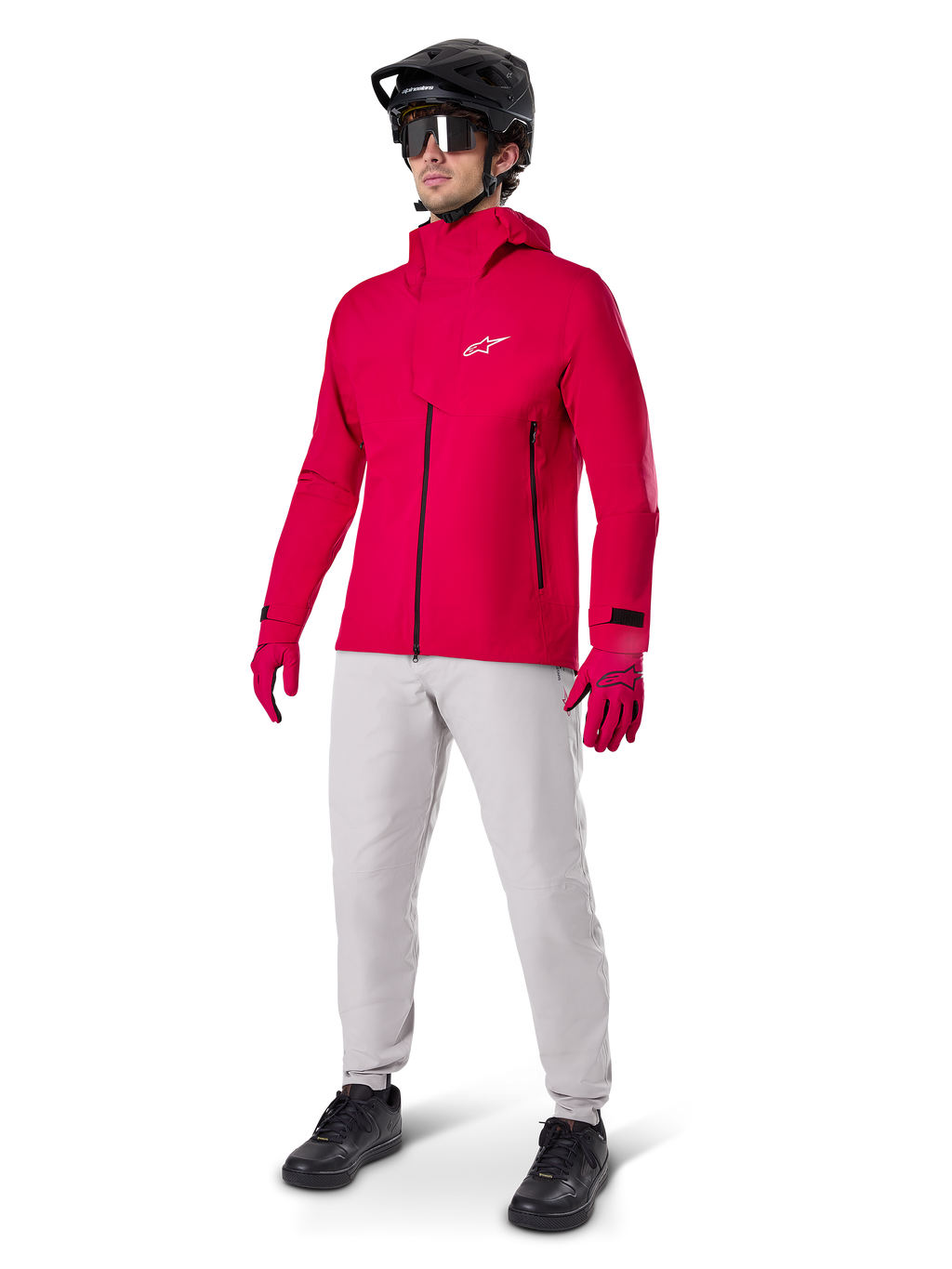 Manlig cyklist i Alpinestars A-Dura Elite vattentät jacka i Red Fluo, kombinerad med ljusgrå tekniska byxor, röda handskar, svart MTB-hjälm med goggles och svarta skor, komplett högpresterande outfit för mountainbike