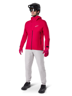 Manlig cyklist i Alpinestars A-Dura Elite vattentät jacka i Red Fluo, kombinerad med ljusgrå tekniska byxor, röda handskar, svart MTB-hjälm med goggles och svarta skor, komplett högpresterande outfit för mountainbike