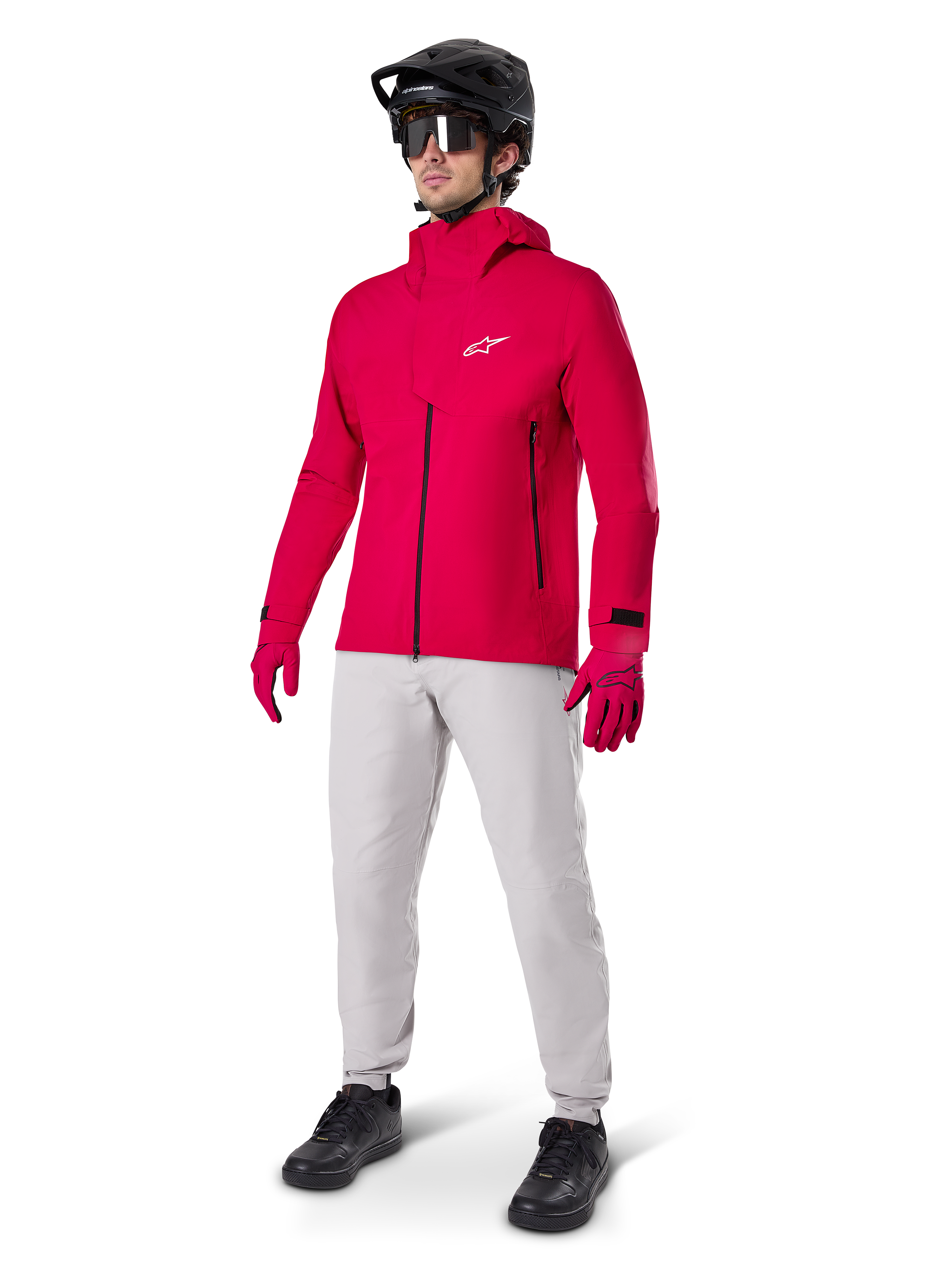 Manlig cyklist i Alpinestars A-Dura Elite vattentät jacka i Red Fluo, kombinerad med ljusgrå tekniska byxor, röda handskar, svart MTB-hjälm med goggles och svarta skor, komplett högpresterande outfit för mountainbike